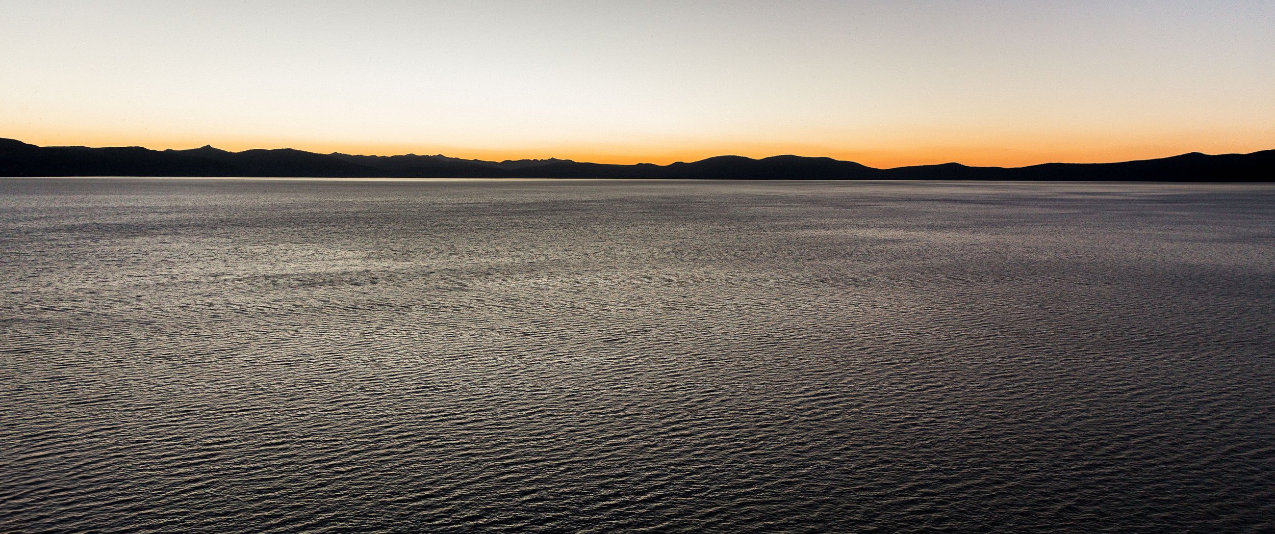 A vast body of water at sunset with distant mountain silhouettes and a clear sky.