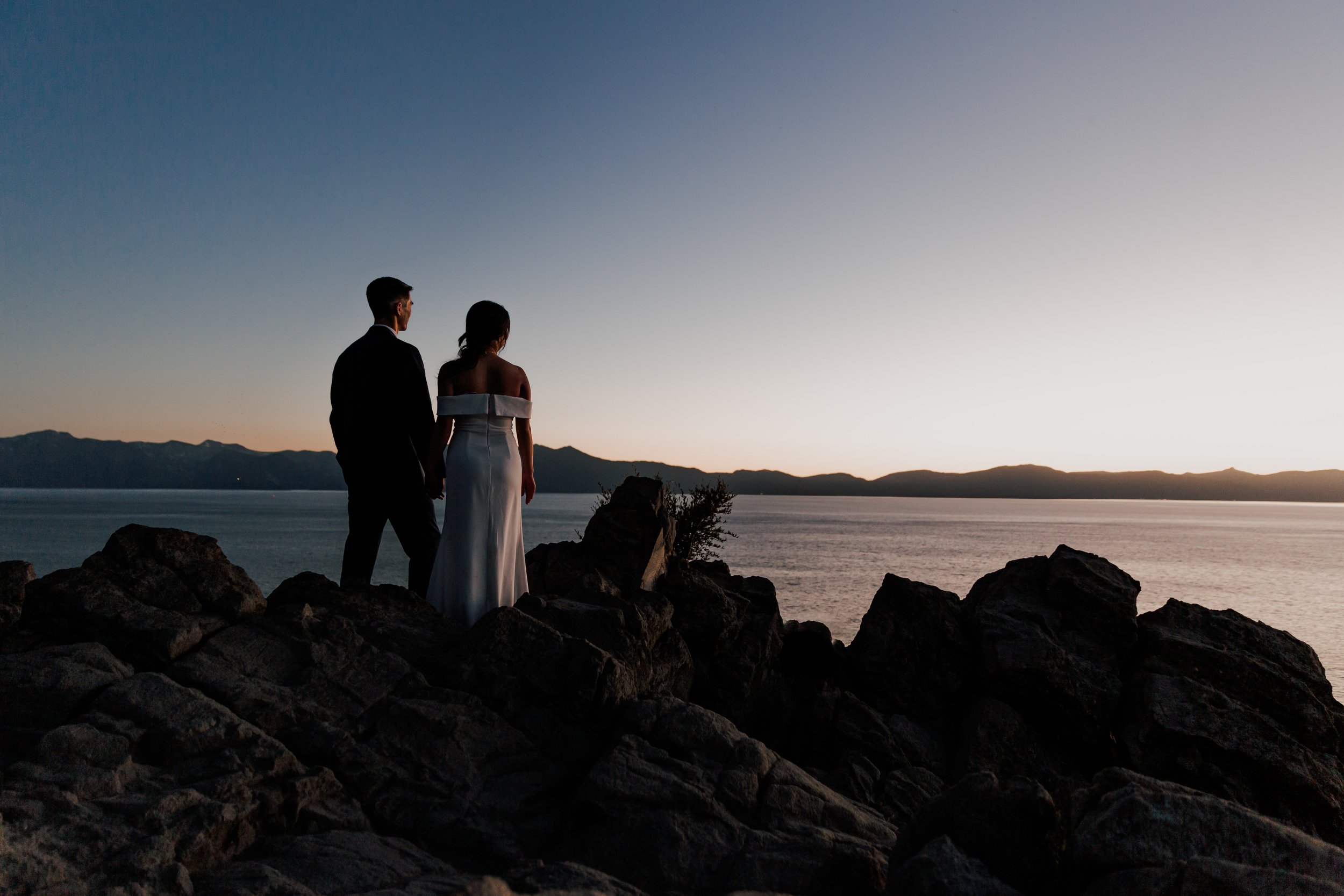 A couple dressed in formal wedding attire standing on rocks by the water during sunset.