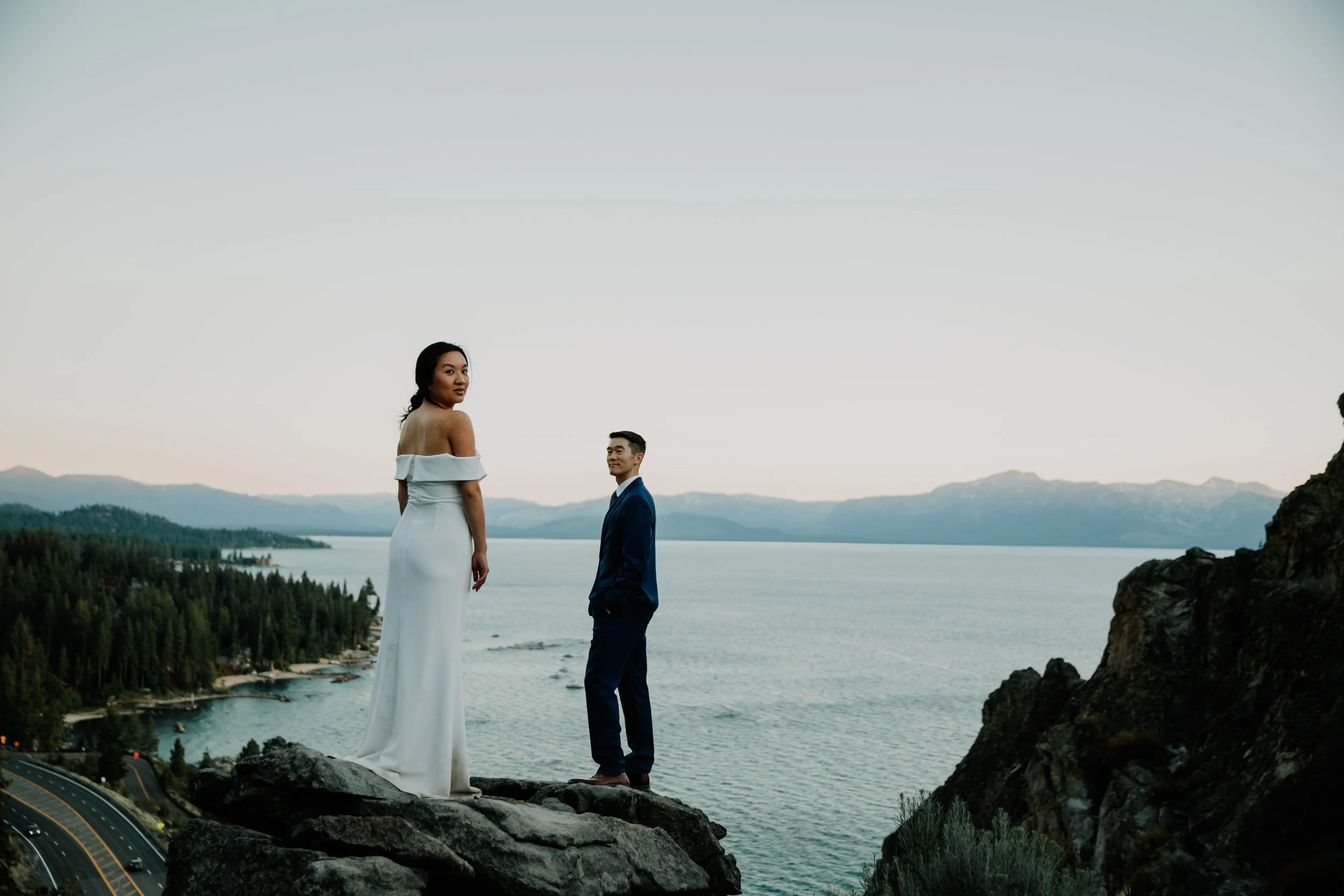 A woman in a white off-shoulder dress and a man in a blue suit standing on rocks overlooking a large lake with mountains in the background during sunset or twilight.
