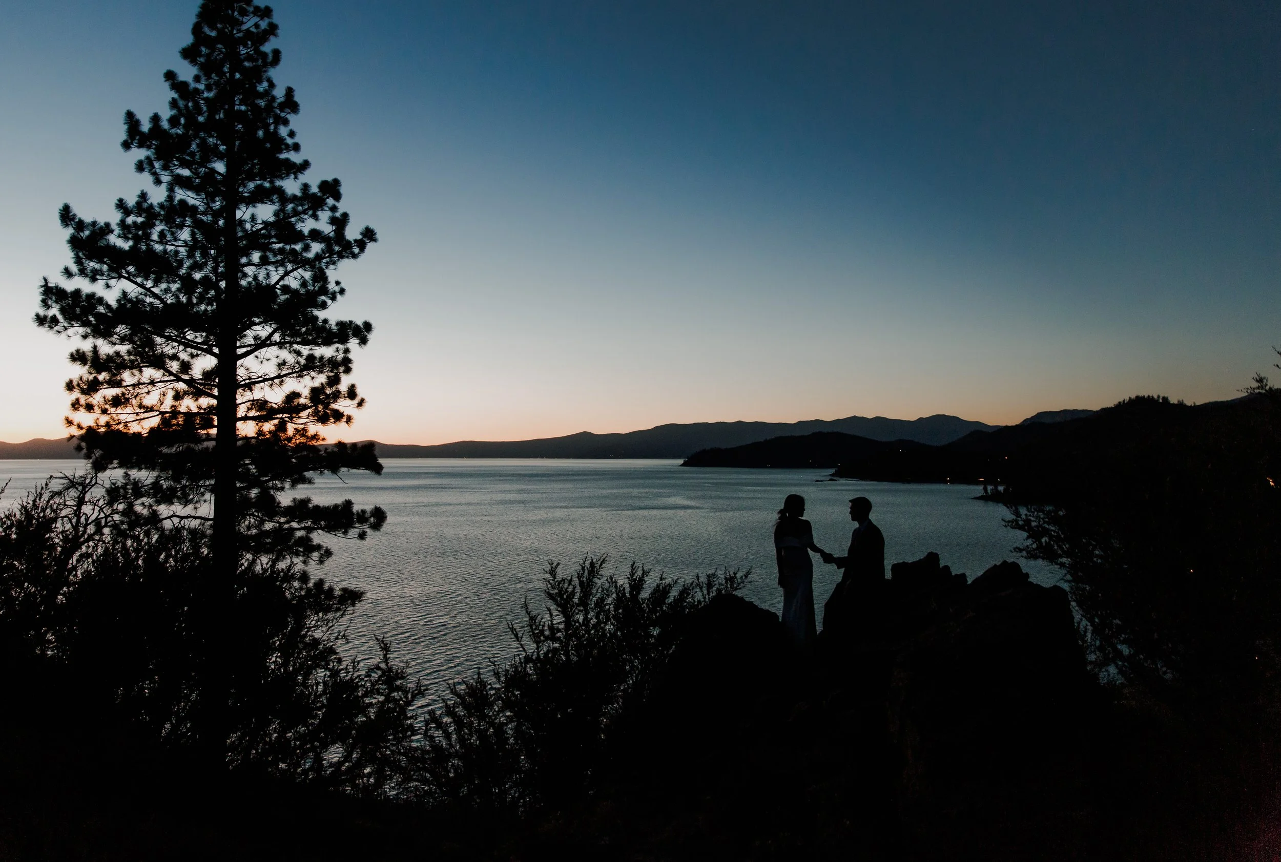 Silhouette of a couple holding hands on rocks by a lake at sunset, with trees and mountains in the background.