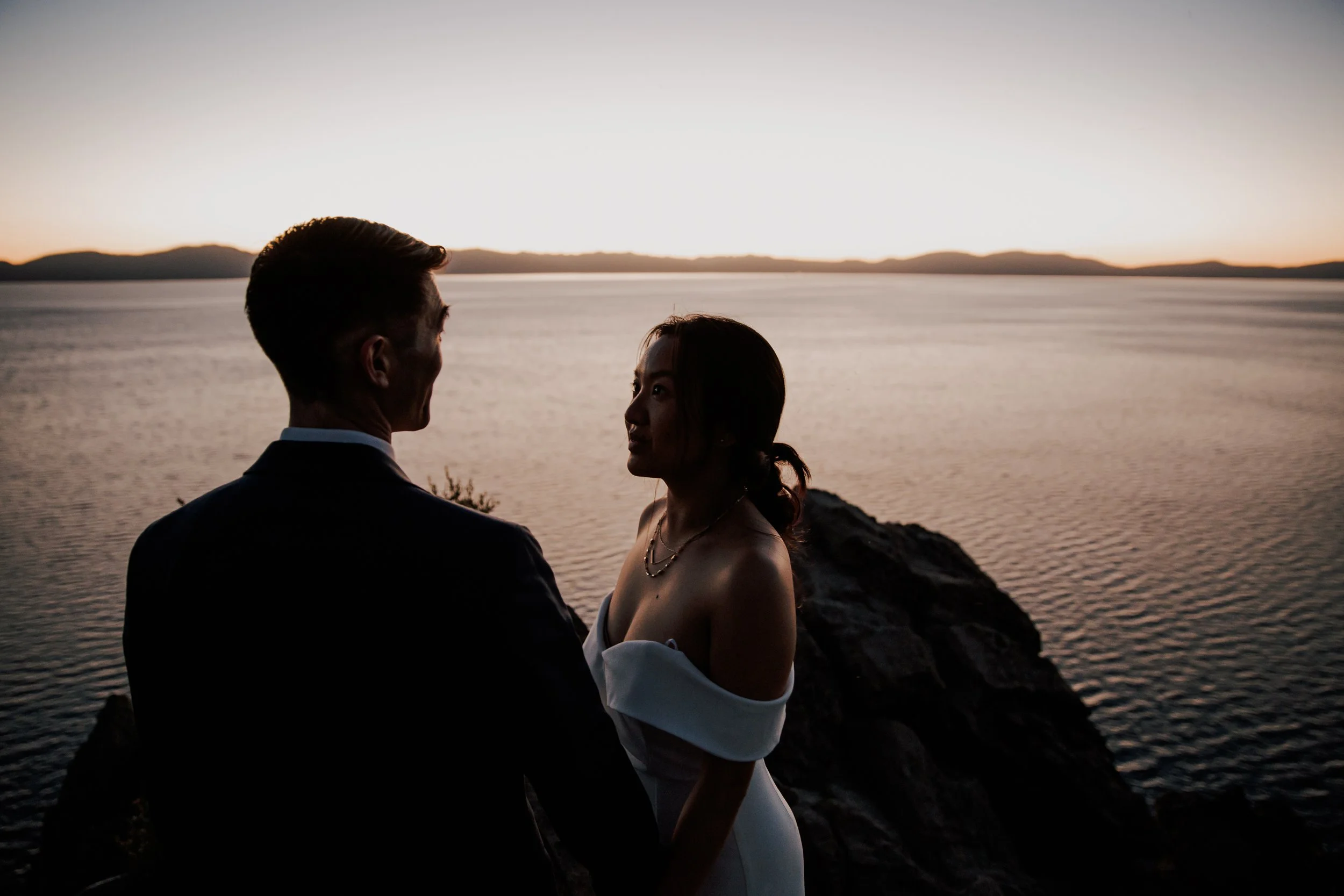 A silhouette of a man and woman facing each other by a body of water during sunset.