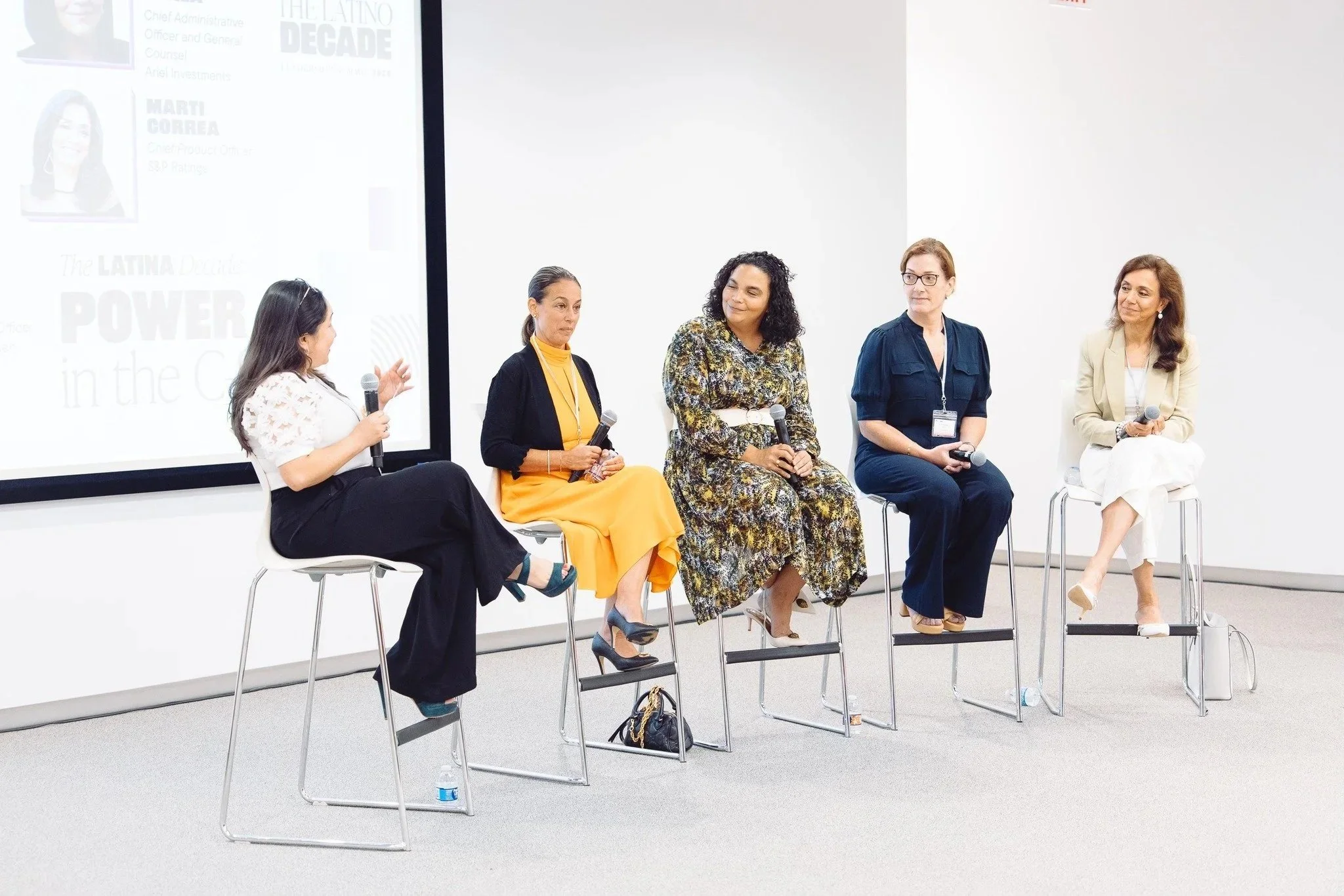 Five women sitting on chairs participating in a panel discussion in front of a large screen, with one woman speaking into a microphone.