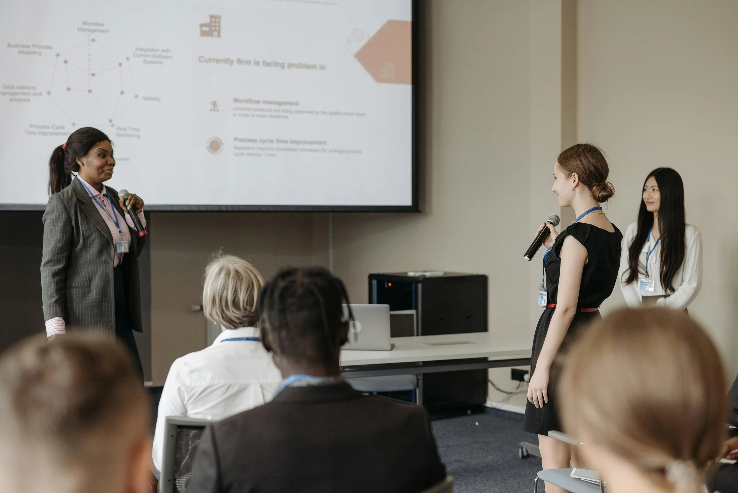 Three women present a business slide in front of an audience at a conference room, with a large screen behind them displaying a presentation slide.