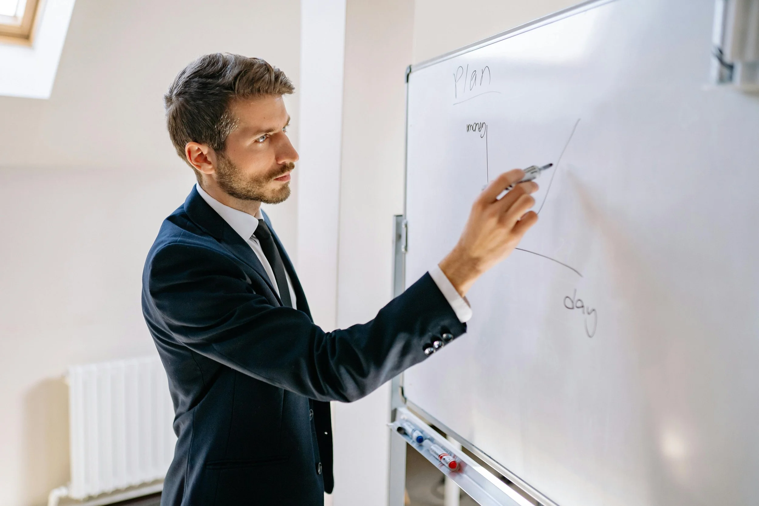 A man in a business suit writing on a whiteboard with a marker in a classroom or office setting.