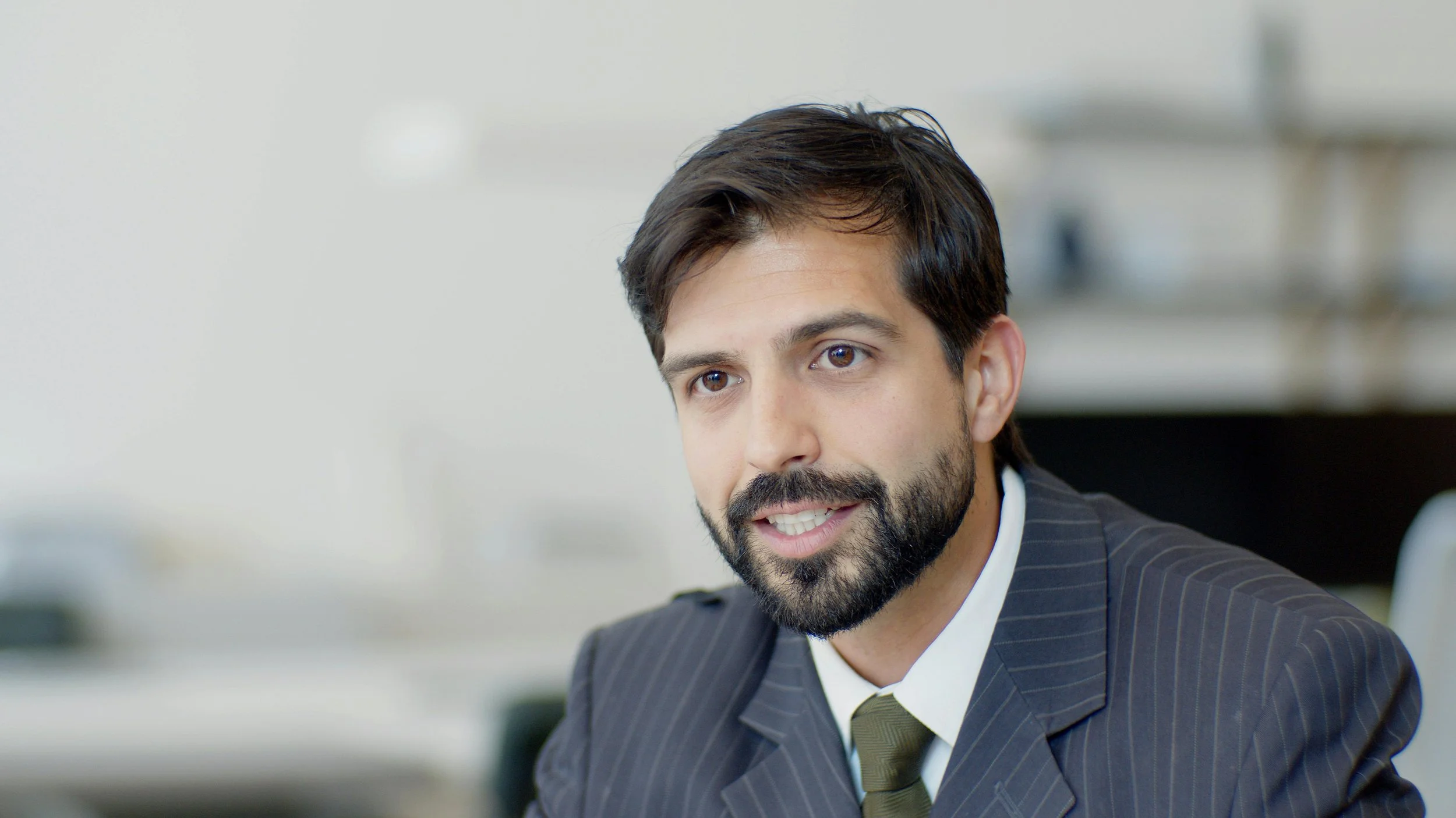 A man with dark hair, a beard, and brown eyes wearing a business suit and striped tie, sitting in an office.