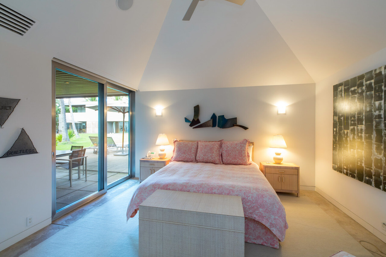  A bedroom in the guest house features a cathedral ceiling to accommodate  a large fan. The painting at right is by Hawaiian artist Mary Mitsuda.  The headboard and bedside tables are from Bielecky Brothers, and the  fabric on the bed is from Quadril