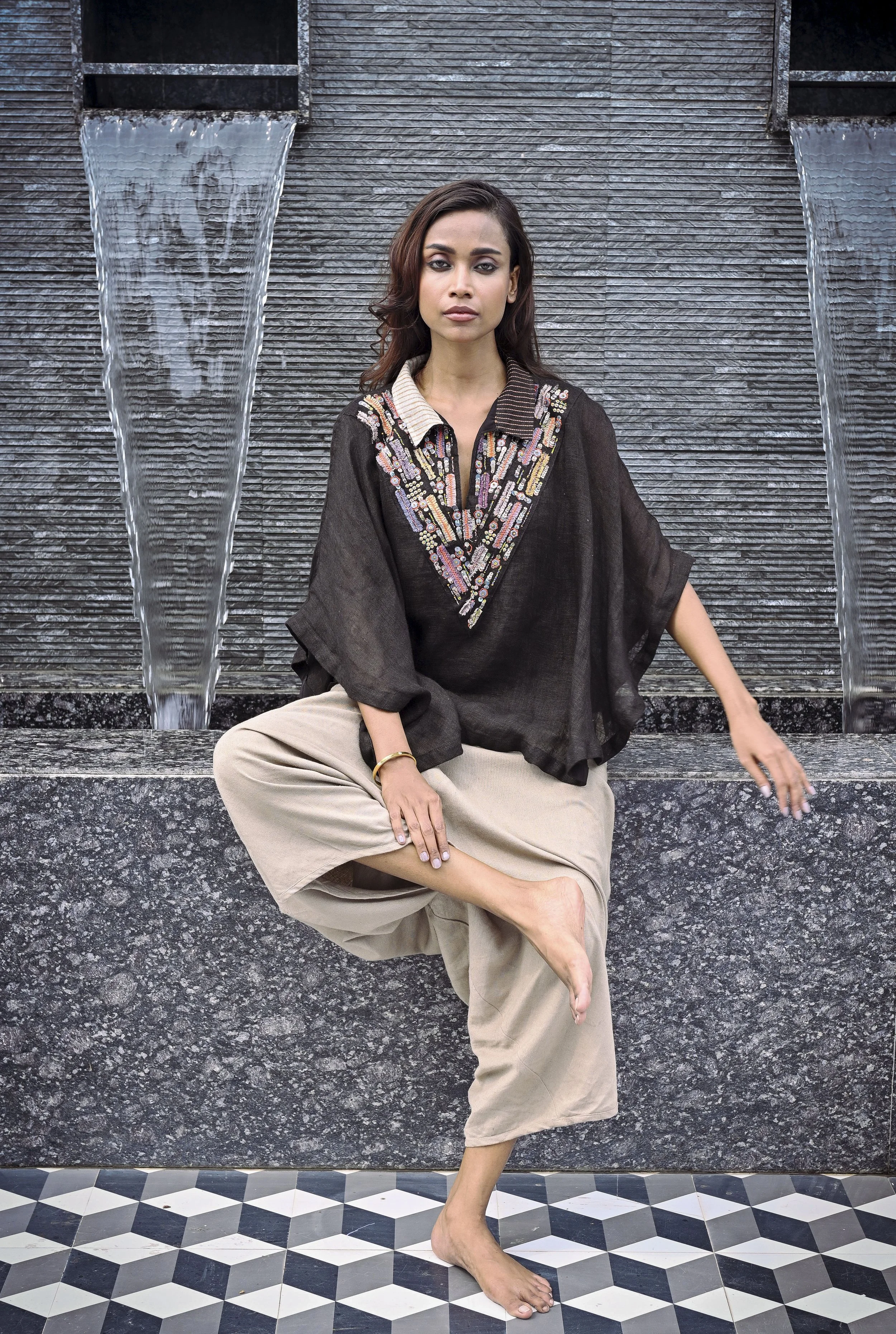 A woman sitting on a stone ledge in front of a water fountain with water cascading down a textured gray wall, wearing a black embroidered blouse and beige wide-leg pants, with one leg crossed and bare feet.