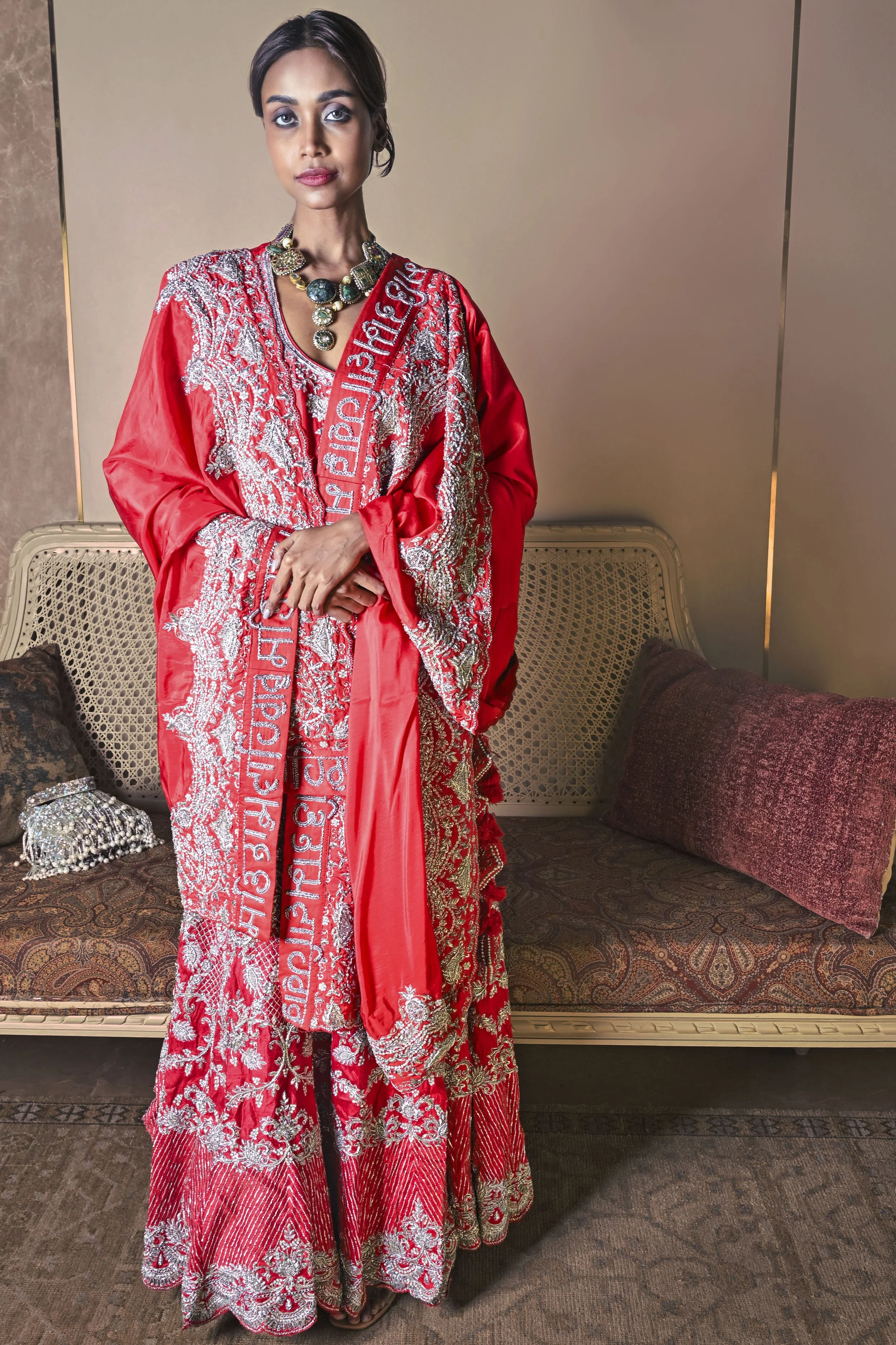 A woman in an elaborate red and silver embroidered traditional dress, accessorized with a statement necklace and sash, standing in a room with a classic style sofa and decorative pillows.