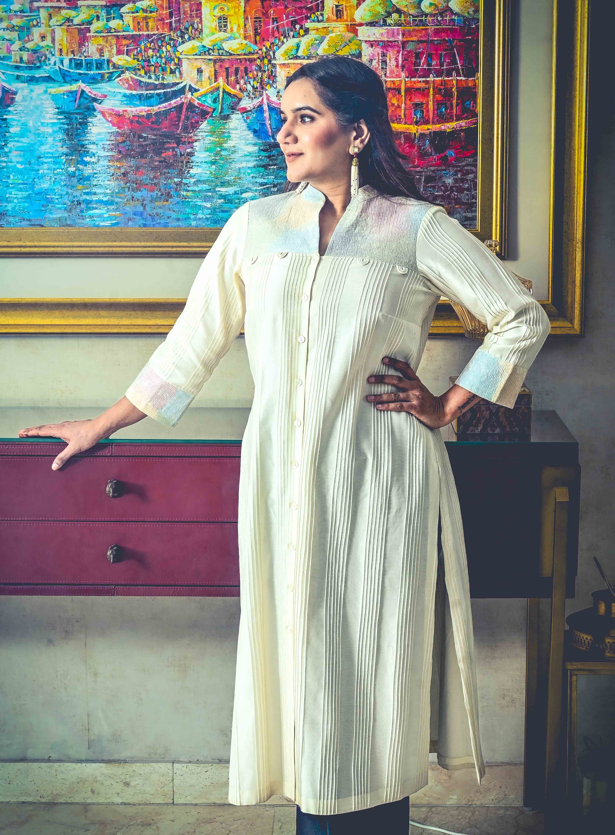 A woman wearing a cream-colored, long, button-down dress with subtle vertical and horizontal pleats, standing indoors in front of a colorful painting of boats on water, a purple dresser, and a gold-framed mirror.