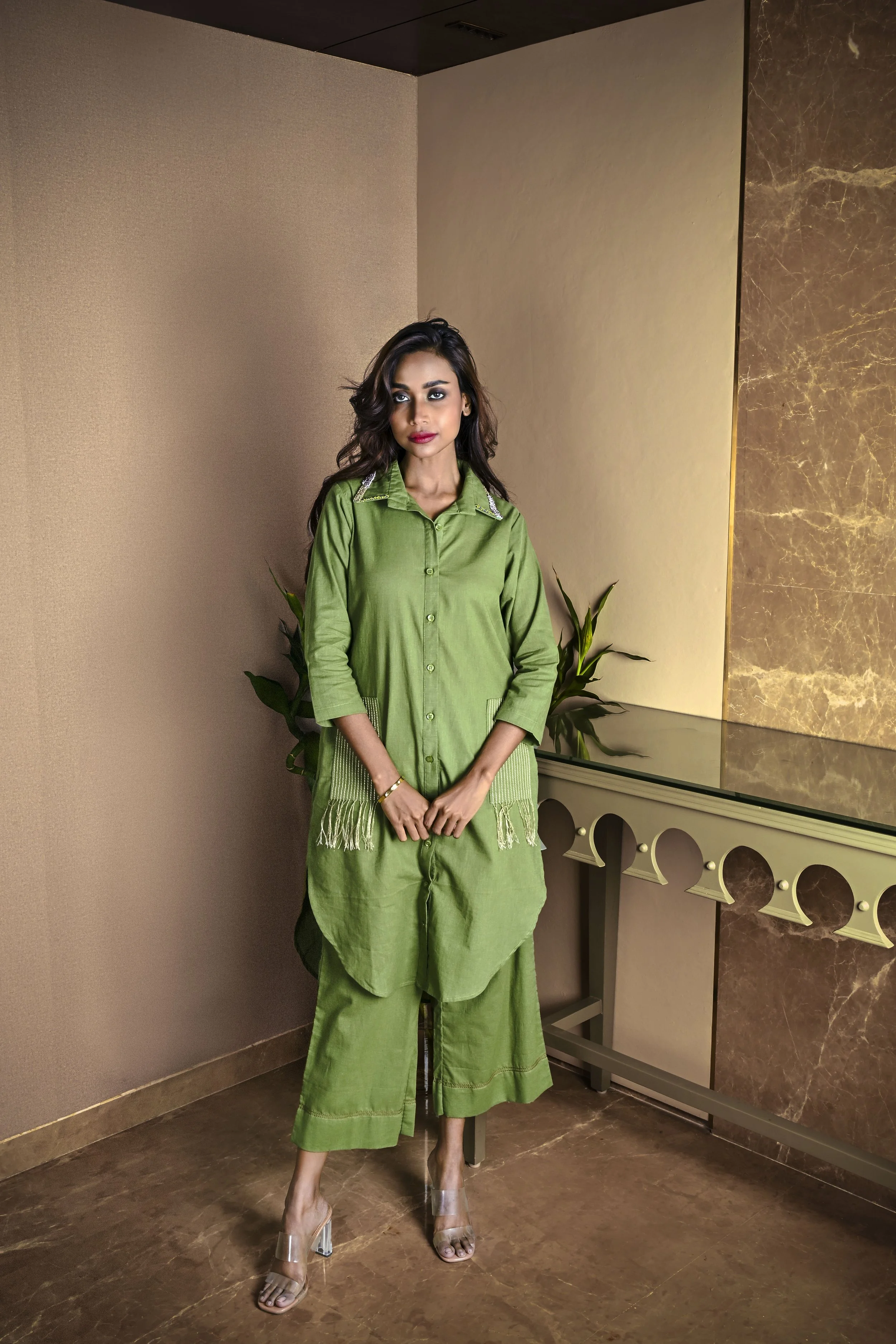 A woman in a green outfit standing indoors near a wall with plants and a marble countertop.