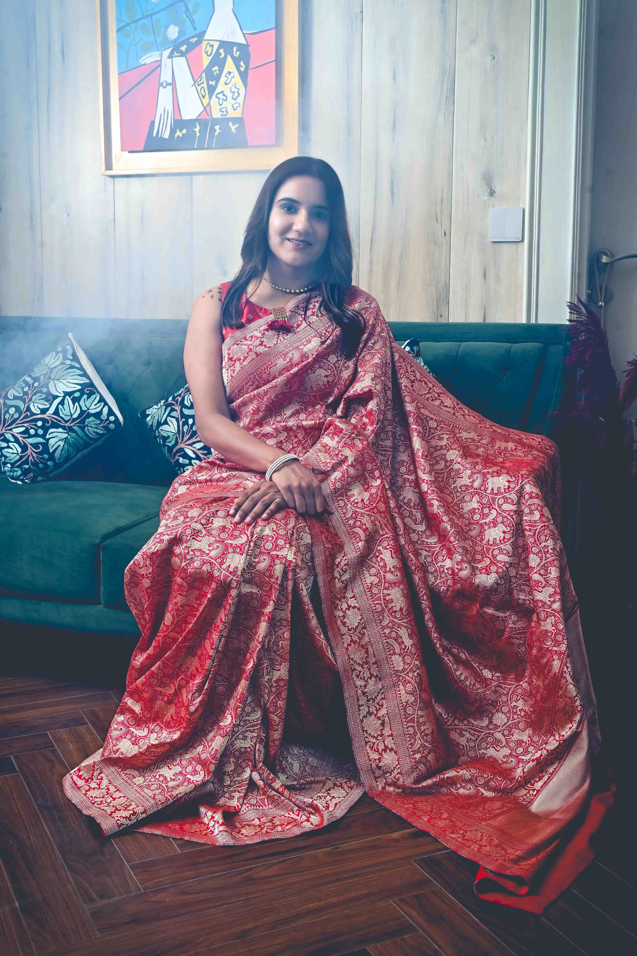 A woman dressed in a traditional red and gold saree sitting on a green couch in a room with wooden panel walls and a colorful framed artwork hanging behind her.