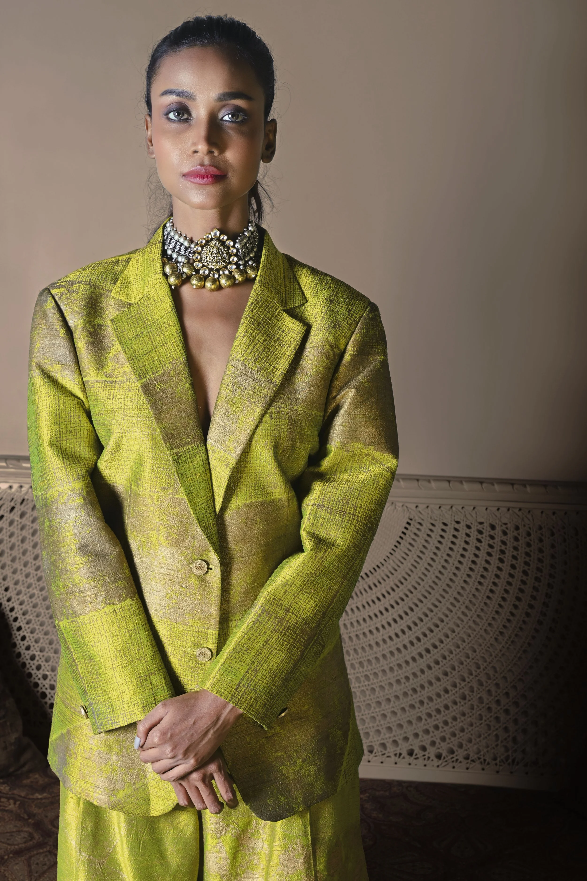 A woman dressed in a bright green patterned blazer and matching skirt, wearing a large pearl and jeweled necklace, standing against a beige wall with a decorative vent.