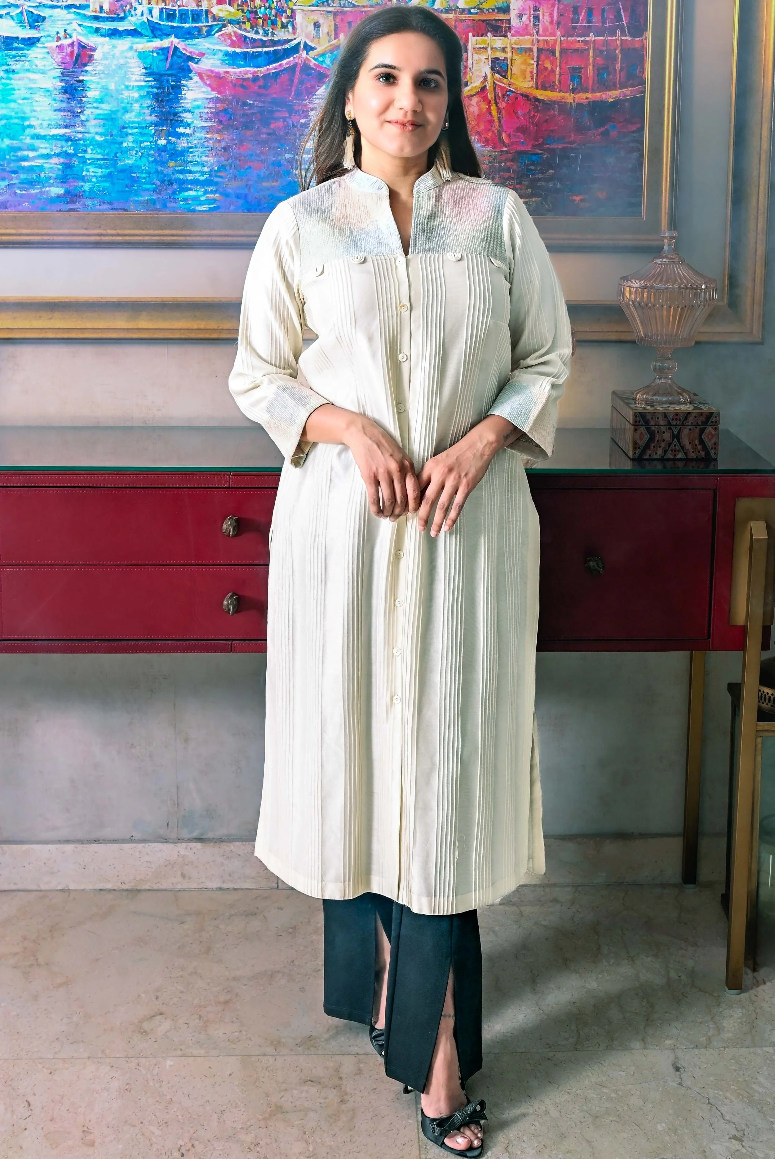 A woman standing in front of a painting, wearing a long, light-colored shirt dress with pleats and black pants with slits, and black heels with bows.