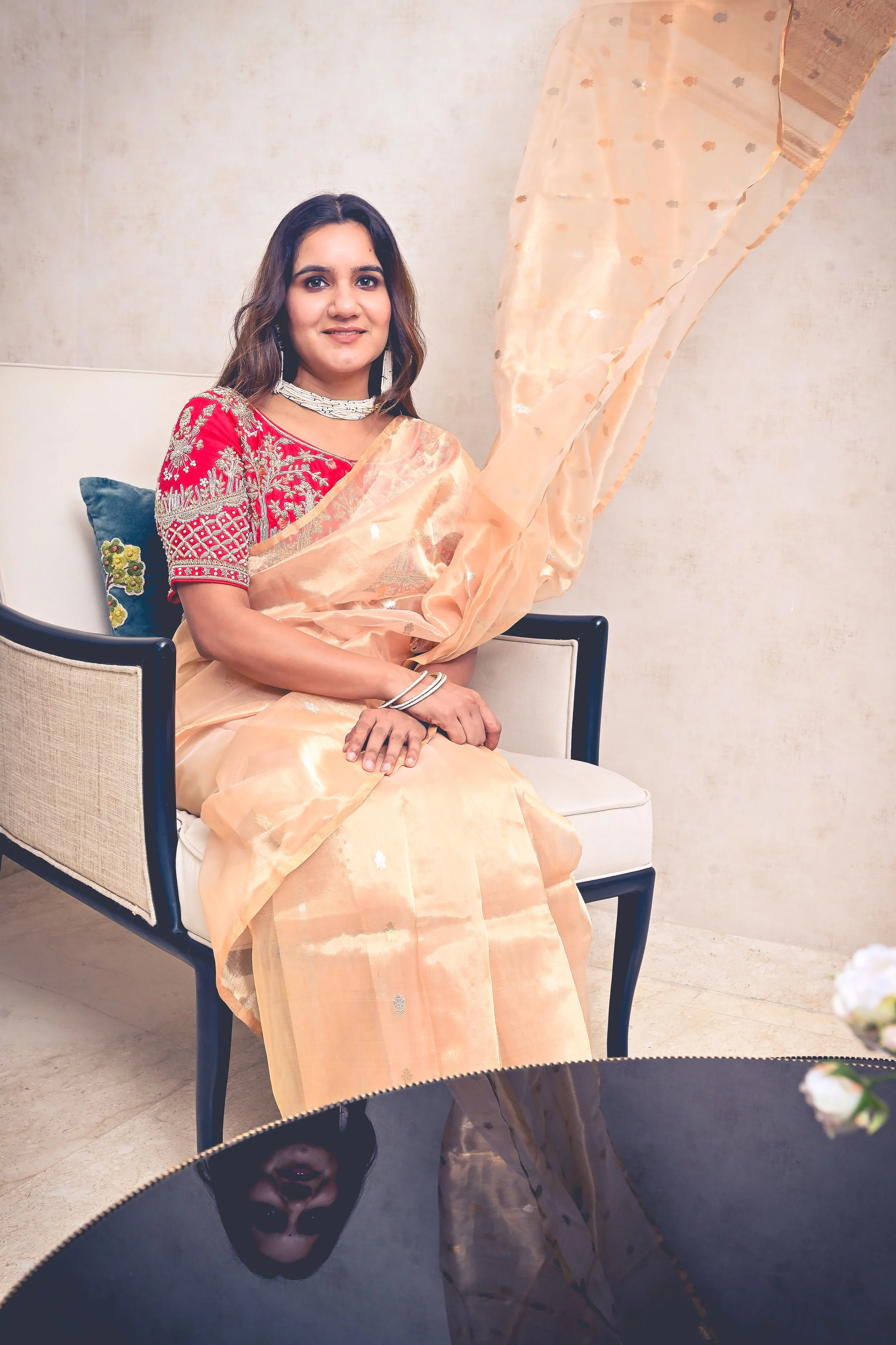 A woman wearing a peach saree with red blouse with intricate embroidery, sitting on a white chair with a beige background and a dark table reflecting her face in the foreground.