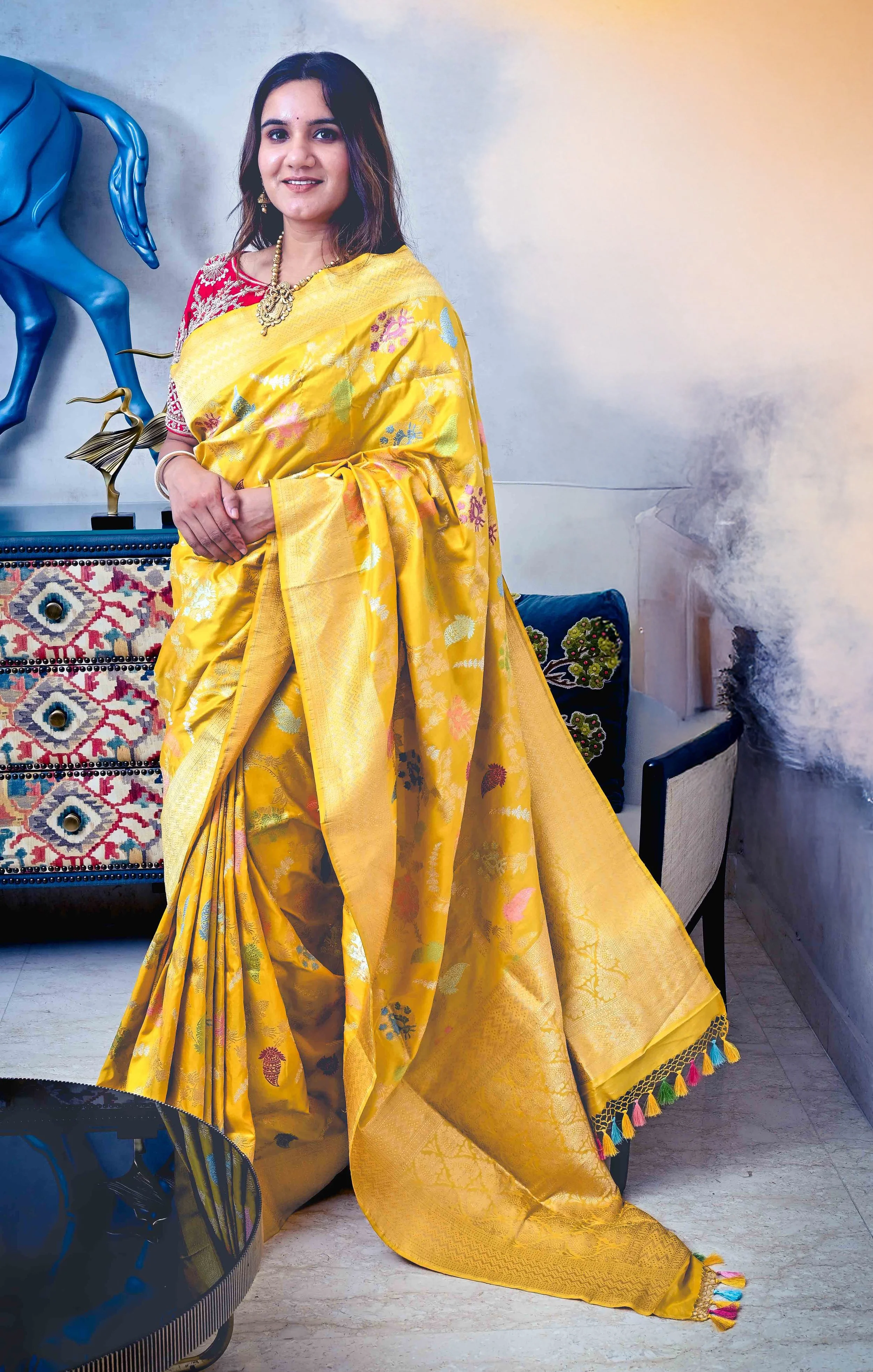 A woman wearing a yellow saree with floral patterns, standing in a stylish room with decorative furniture and artwork.