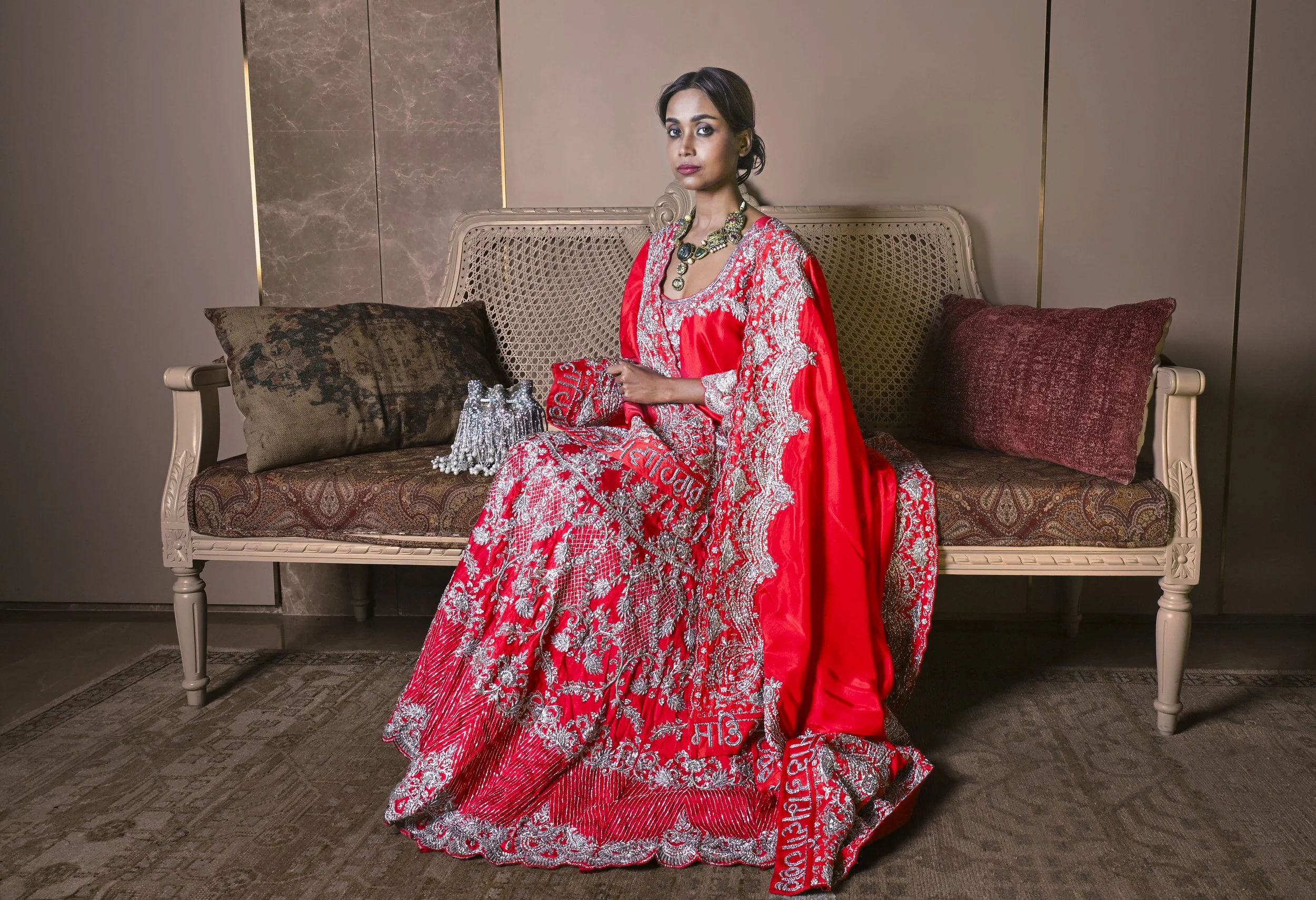 A woman dressed in a red and silver traditional South Asian outfit sits on a vintage-style cream-colored bench with brown and red cushions in a luxurious indoor setting.