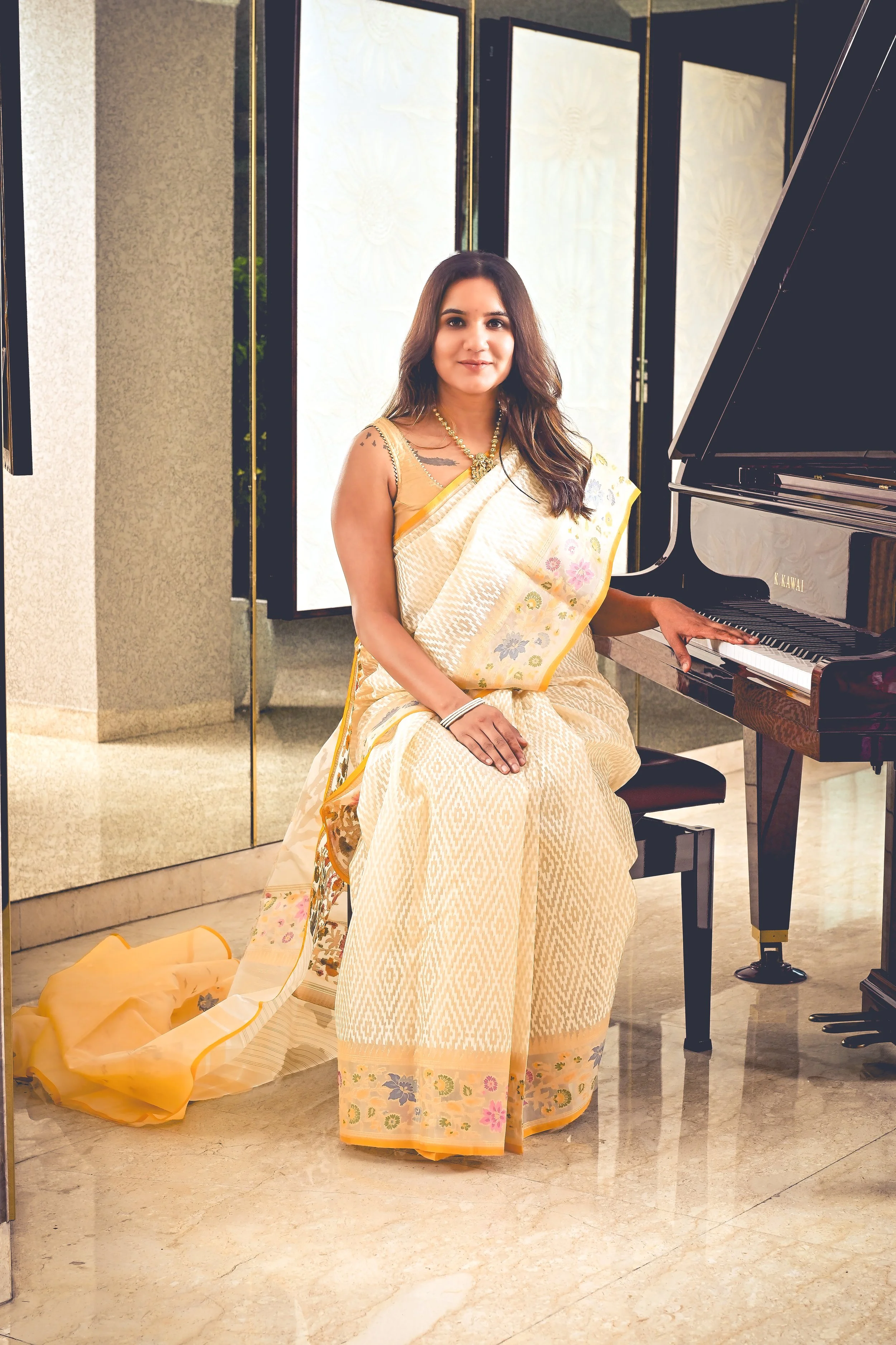 Woman in a traditional light-colored saree sitting at a black grand piano.