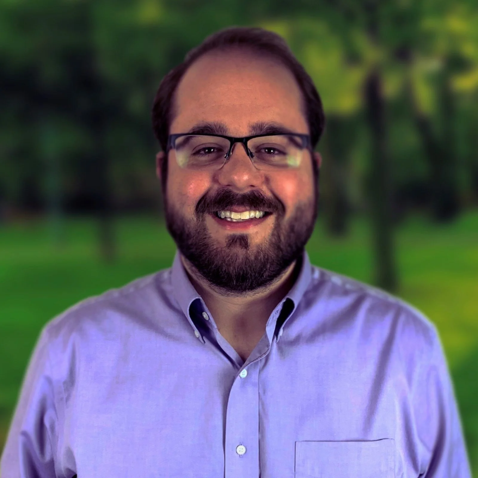 A man with glasses and a beard smiling outdoors in front of a blurred green background.