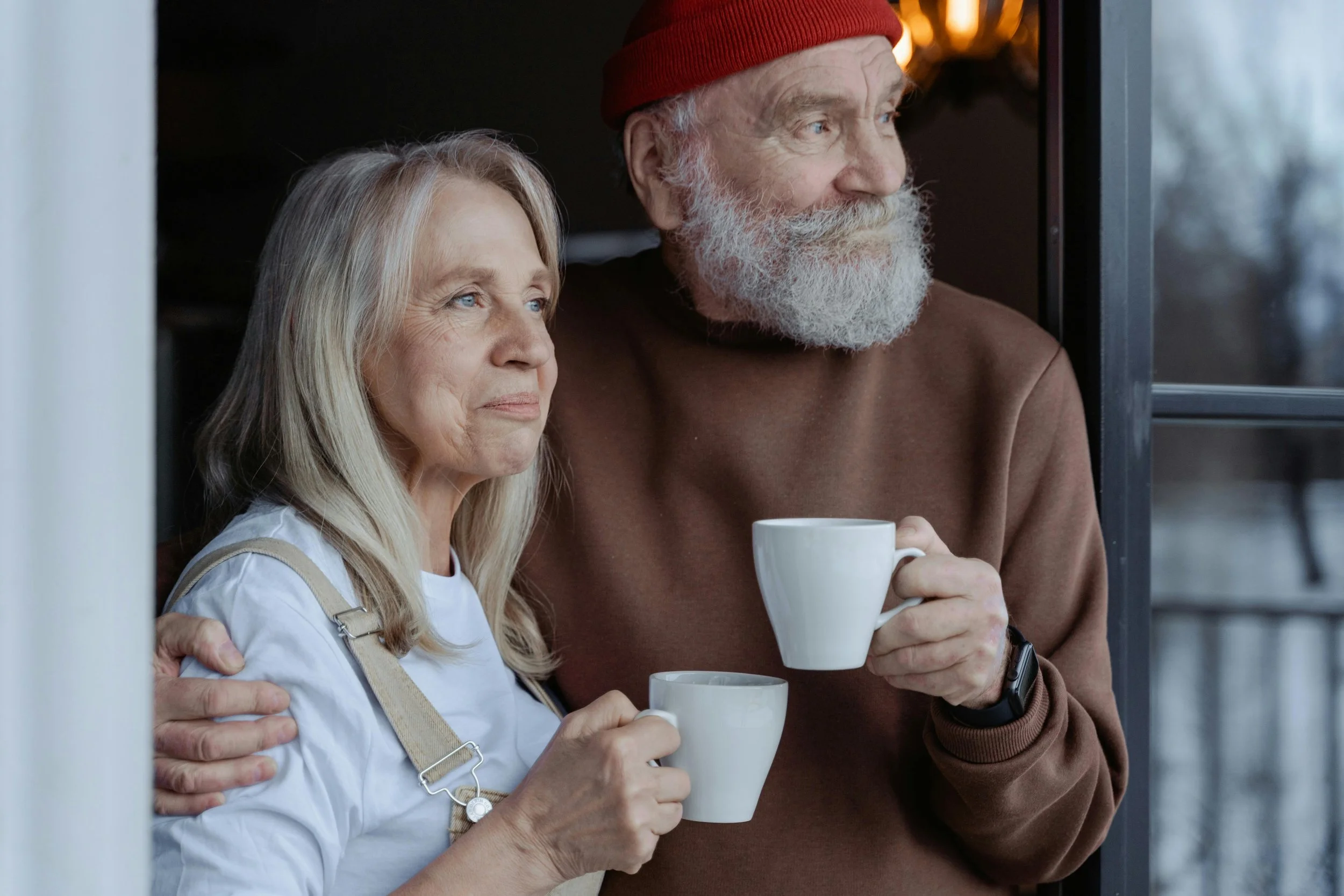An elderly couple standing on a porch, holding white mugs and gazing outside, with the man's arm around the woman's shoulder.