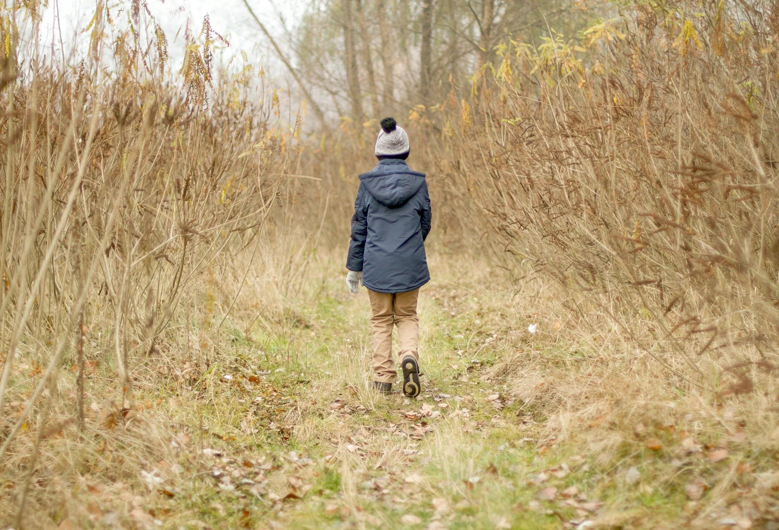 Person walking down a dirt trail through a wooded area with bare trees and brown grass, wearing a blue jacket, tan pants, and a gray knit hat with a pom-pom.