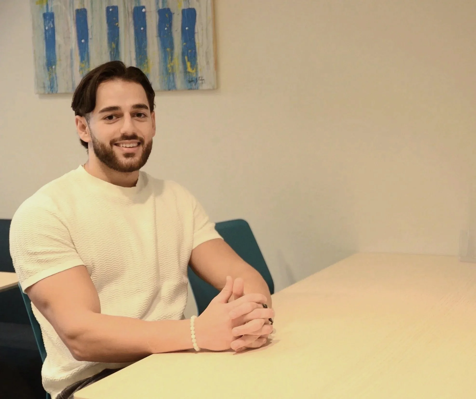 En man sitter vid ett bord i ett rum med beige väggar och ett abstrakt konstverk. Han har mörkt, vågigt hår och skägg, och bär en ljus tröja.