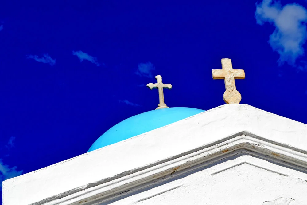 White church rooftop with two crosses on top, one on a blue dome, against a blue sky with scattered clouds. Greece travel photography prints for sale. 