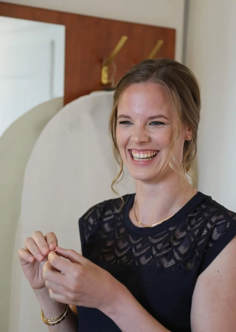 Lächelnde Frau mit braunen, im Nacken hochgesteckten Haaren, trägt ein schwarzes Top mit Spitze, goldene Armbänder und eine Halskette, im Hintergrund ein Kopfteil eines Bettes mit Nachttischlampe.