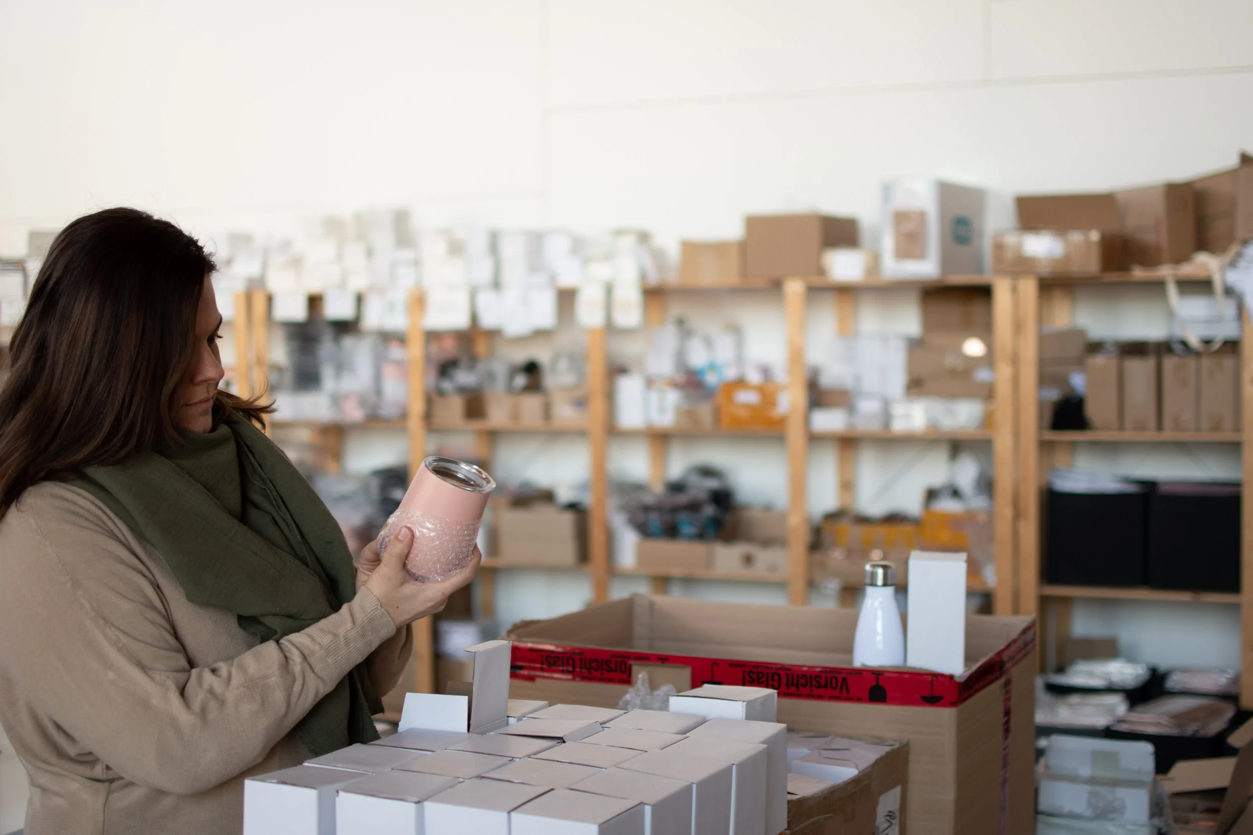 Frau in einem Lagerraum hält einen Gegenstand in der Hand, umgeben von Kartons und Regalen.