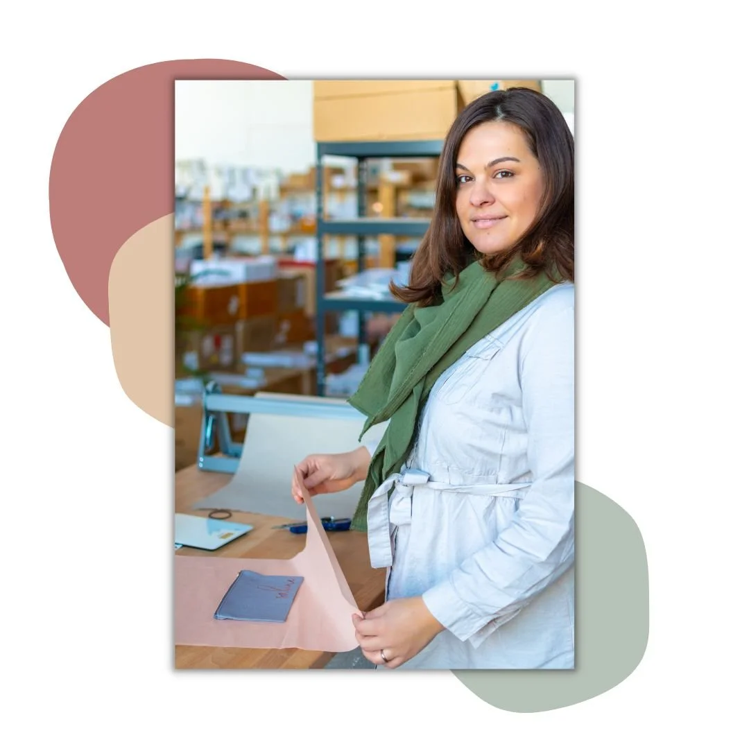 Eine Frau mit einem grünen Schal steht an einem Tisch in einem Lager oder Büro und hält ein Blatt Papier. Im Hintergrund sind Regale mit Kartons und Büromaterial zu sehen.