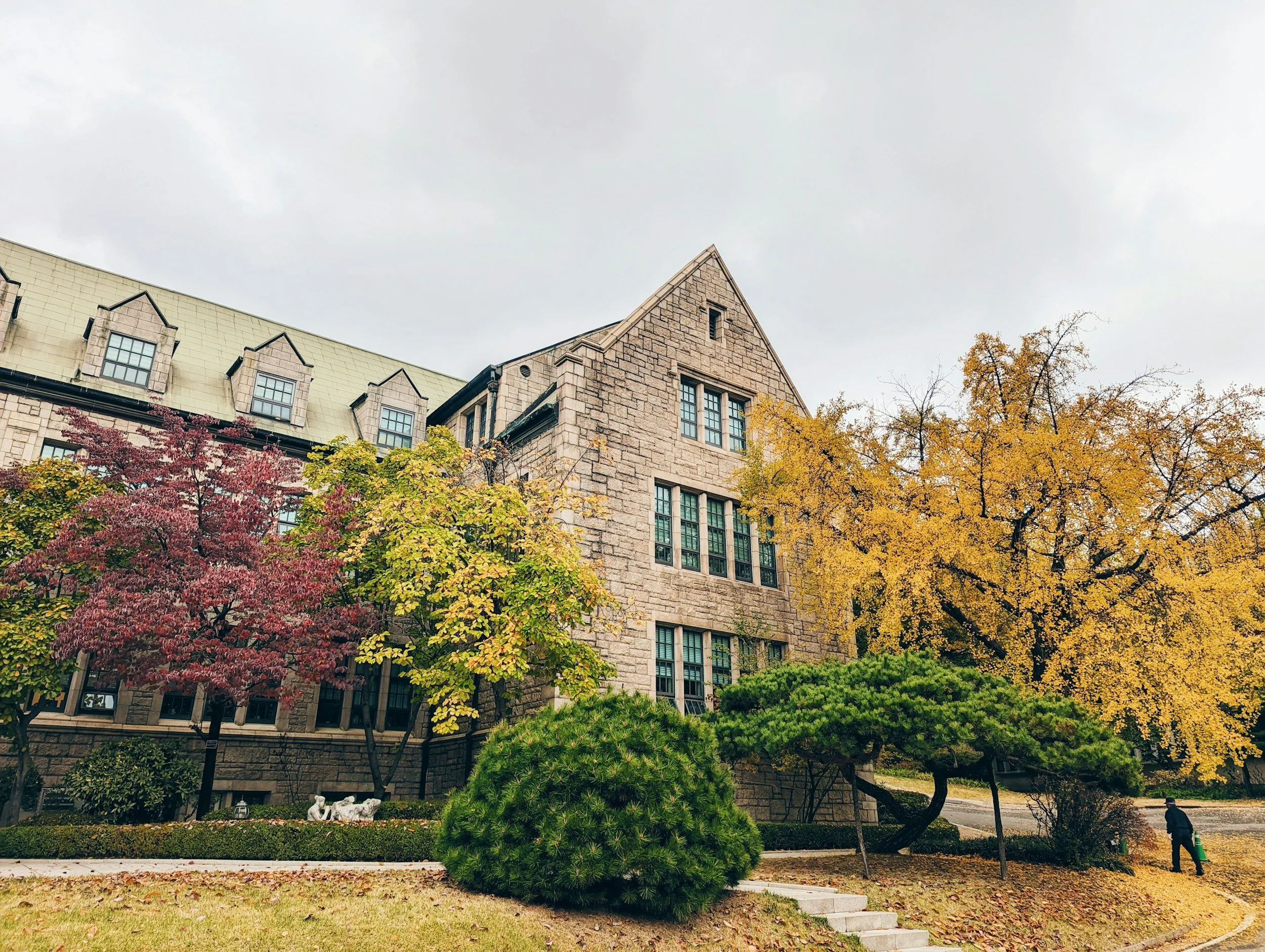 A stone building with multiple windows and a sloped roof, surrounded by colorful autumn trees and shrubs, with a person walking on the sidewalk.