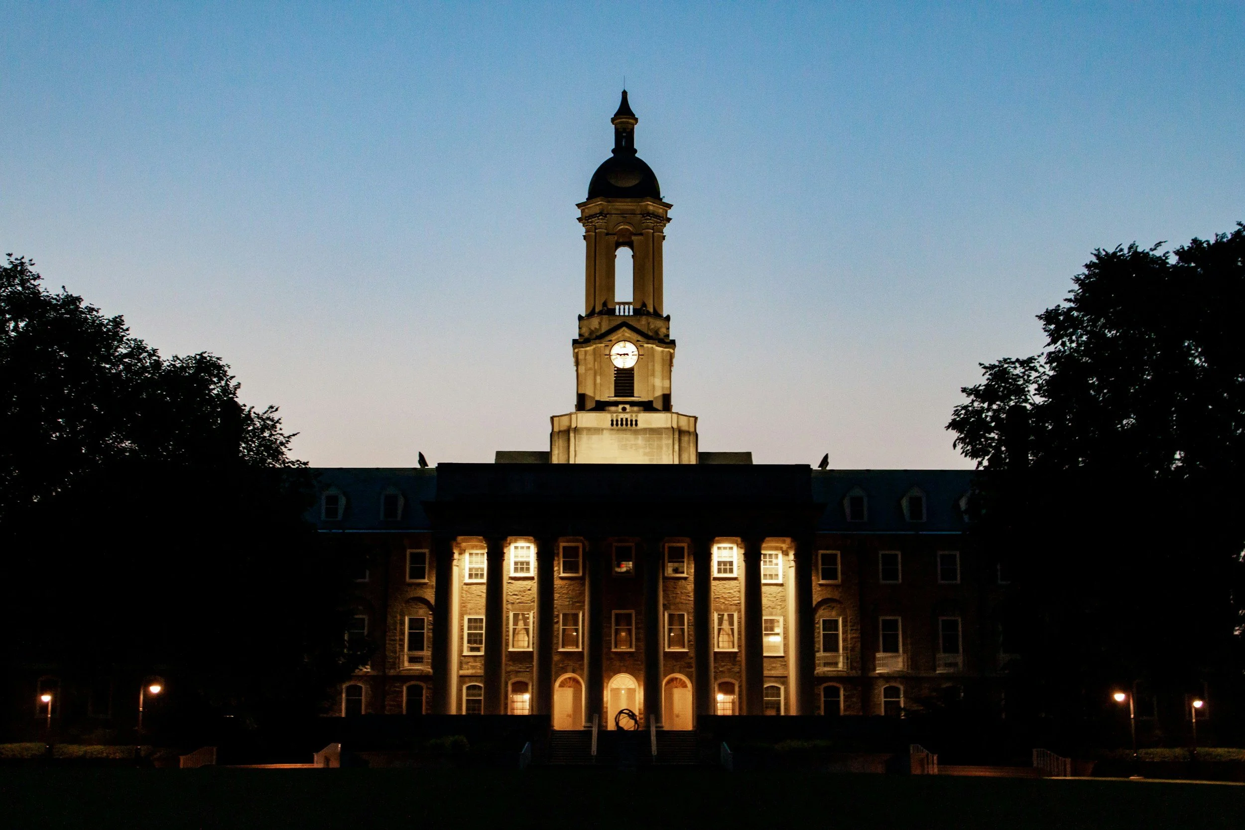 Nighttime view of a historic government building with a clock tower, illuminated by exterior lights, flanked by trees.