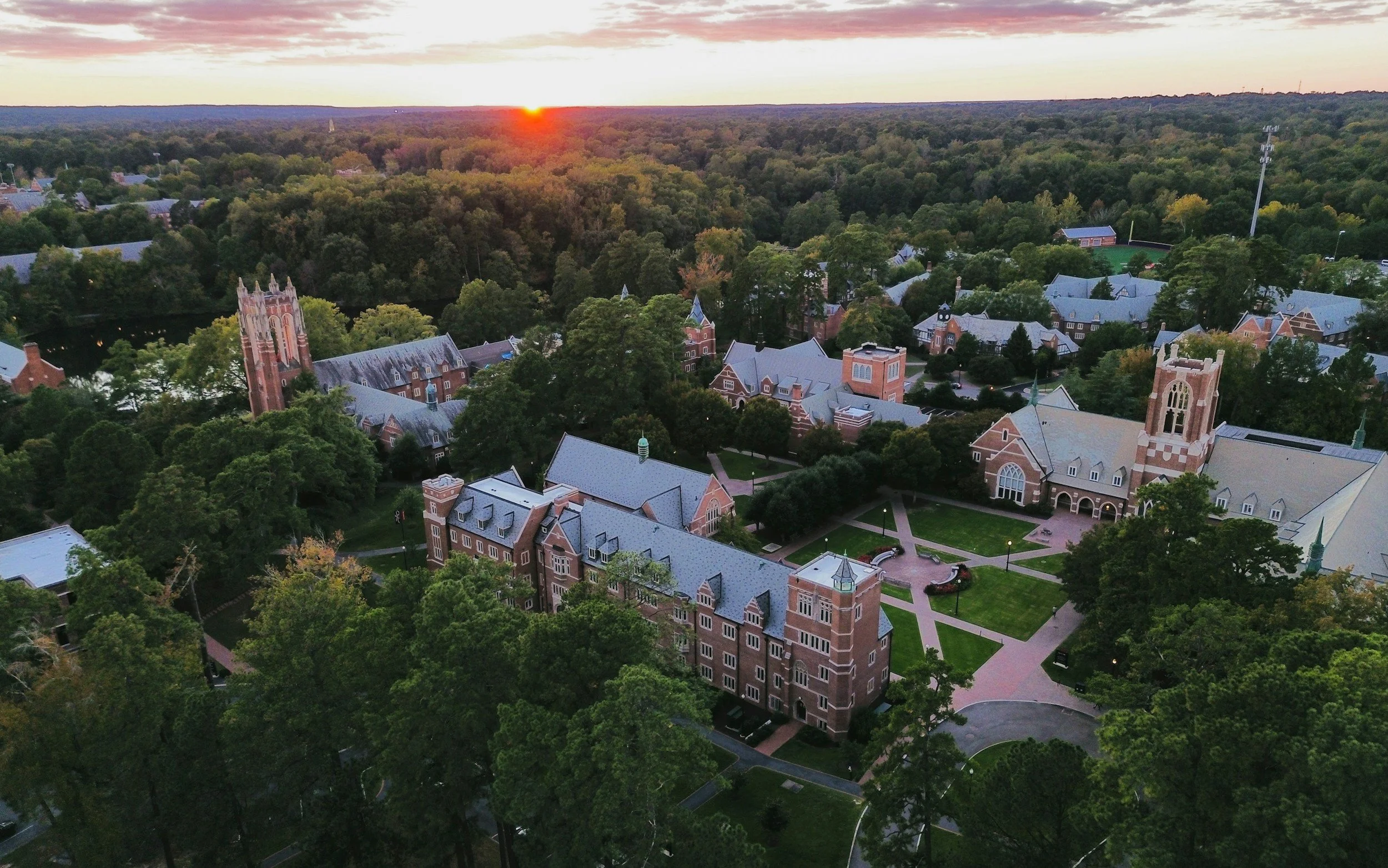 Aerial view of a university campus at sunset, featuring historic brick buildings, lush green trees, and a central courtyard with walkways.