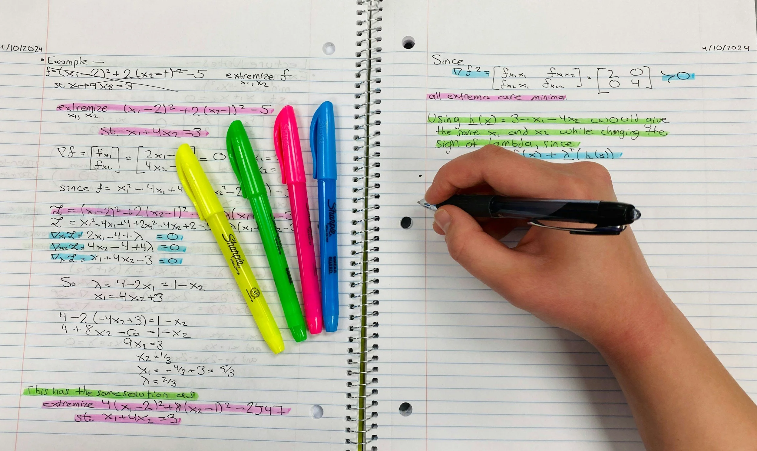 Open spiral notebook pages filled with handwritten mathematical equations and notes, four colorful highlighters (yellow, green, pink, and blue) resting on the left page, and a hand writing with a black pen on the right page.
