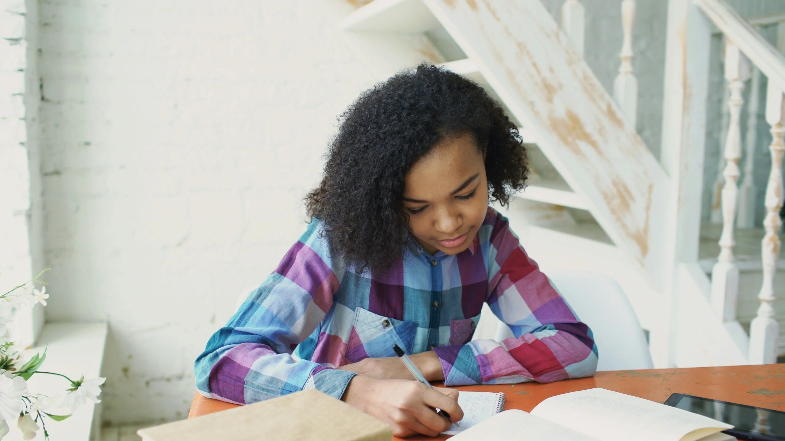 A young girl with curly dark hair writing in a notebook at a wooden table, with books and a tablet nearby, in a bright room with stairs and a white brick wall in the background.