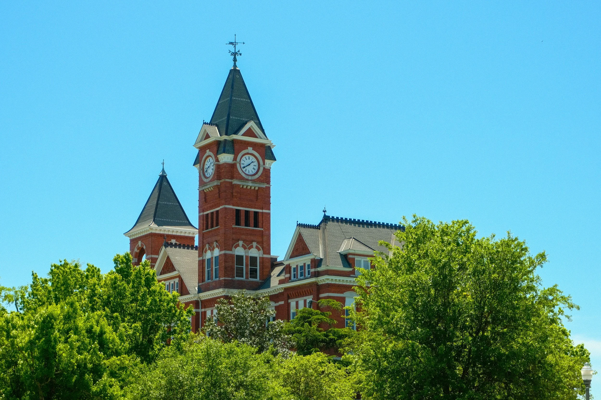A historic red brick courthouse with a clock tower, surrounded by green trees and a clear blue sky.