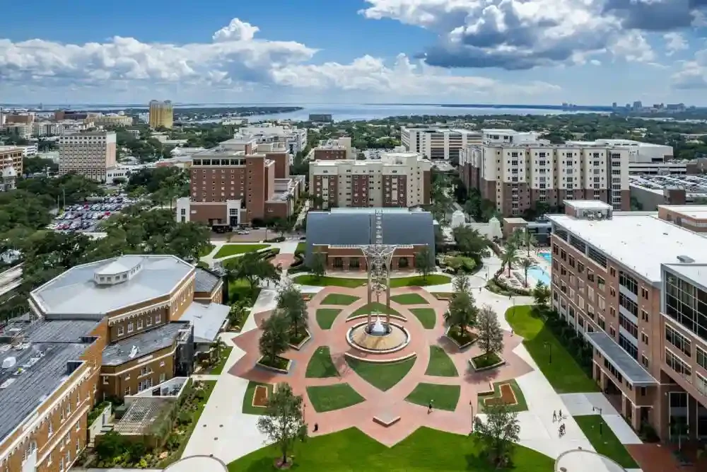 Aerial view of a cityscape with a park featuring a fountain, walking paths, trees, and surrounding buildings under a partly cloudy sky.