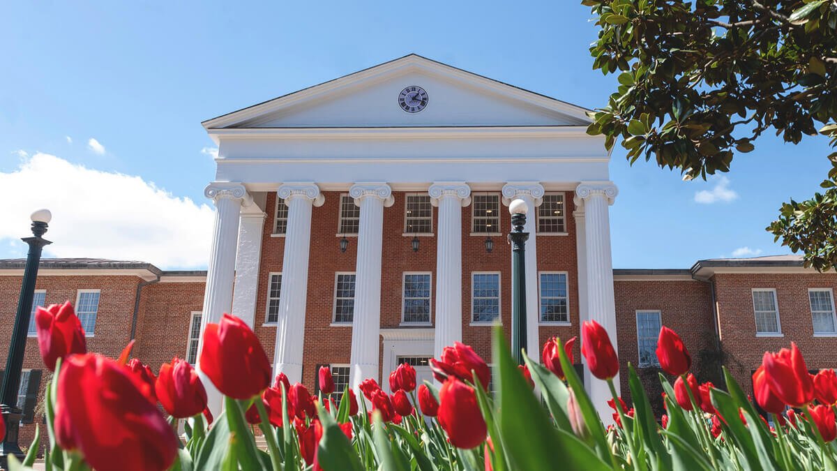 A university or college building with white columns, brick walls, and a clock at the top of the facade, surrounded by red tulips and green trees under a blue sky.