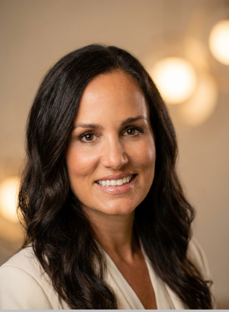 A woman with long dark hair, wearing a light-colored blazer, smiling in front of a blurred background with warm lights.