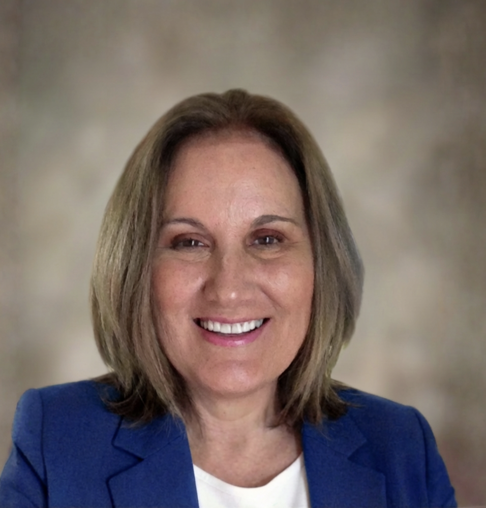 A woman with shoulder-length brown hair smiling, wearing a blue blazer and a white top, against a blurred neutral background.