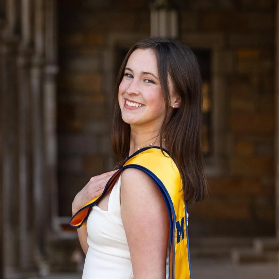 Young woman with brown hair smiling at the camera, wearing a white dress and a yellow sash, outdoors.