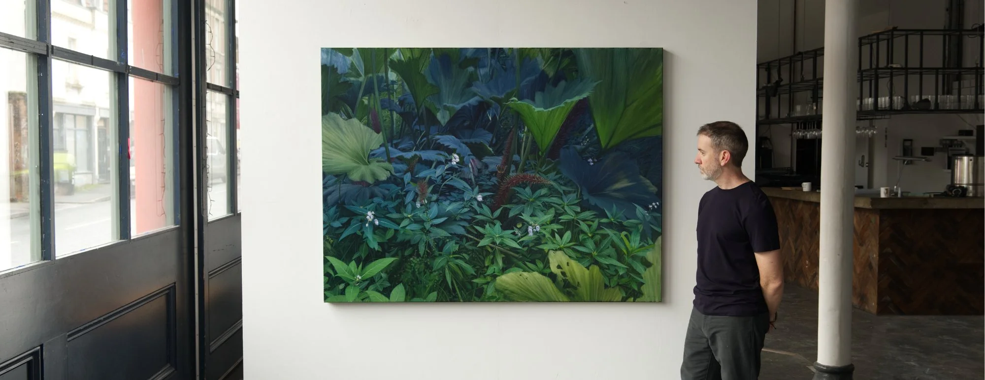 A man with a beard wearing a black t-shirt stands and looks at a large painting of lush green jungle plants inside an art gallery.