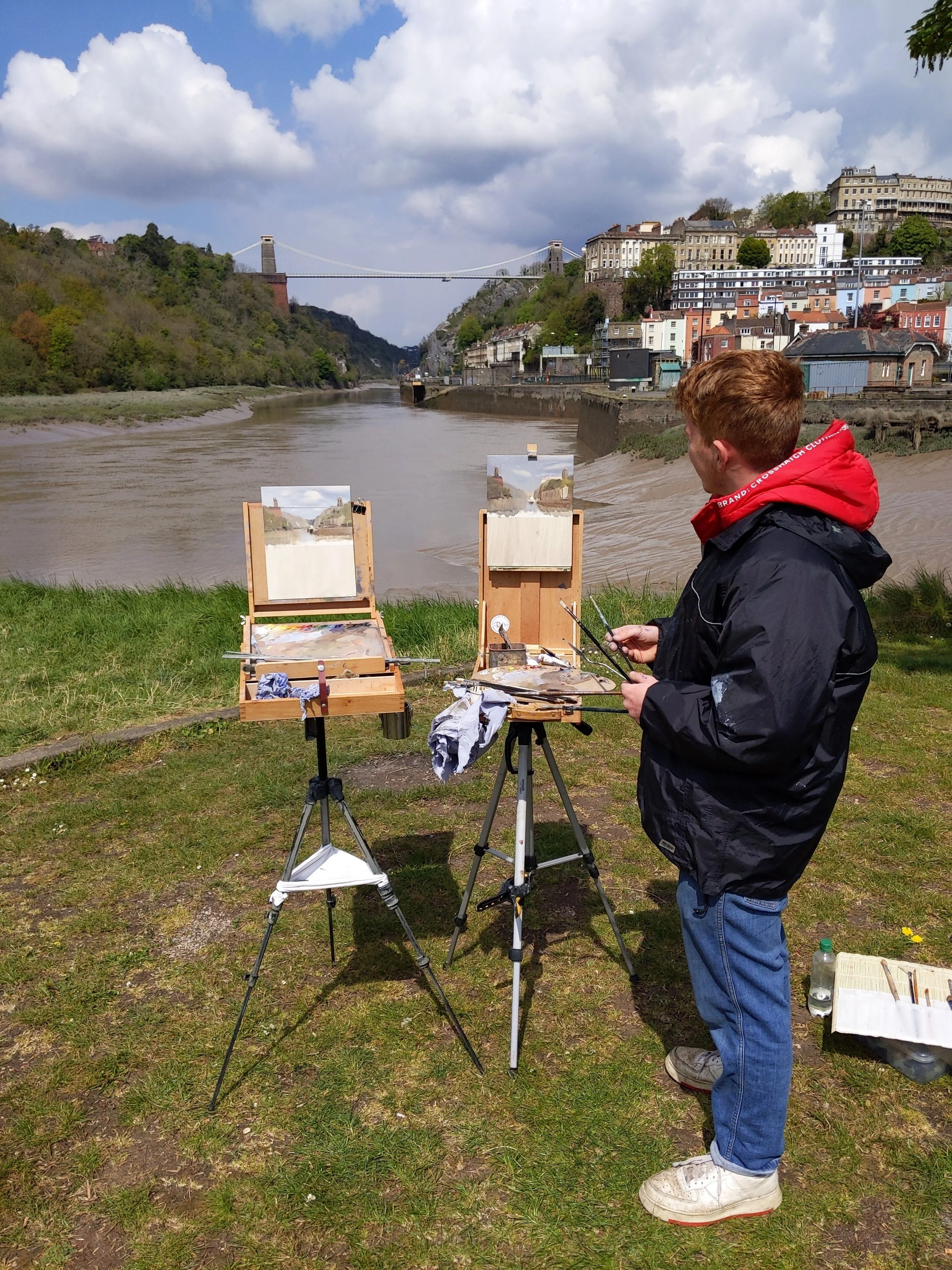 A person with reddish hair wearing a black jacket and blue jeans stands outdoors near the riverbank, holding a paintbrush and palette. Two small easels with paintings are set up in front of him. The river flows through a cityscape with colorful buildings on the hillside and a bridge in the background. The sky is partly cloudy with large clouds and patches of blue.