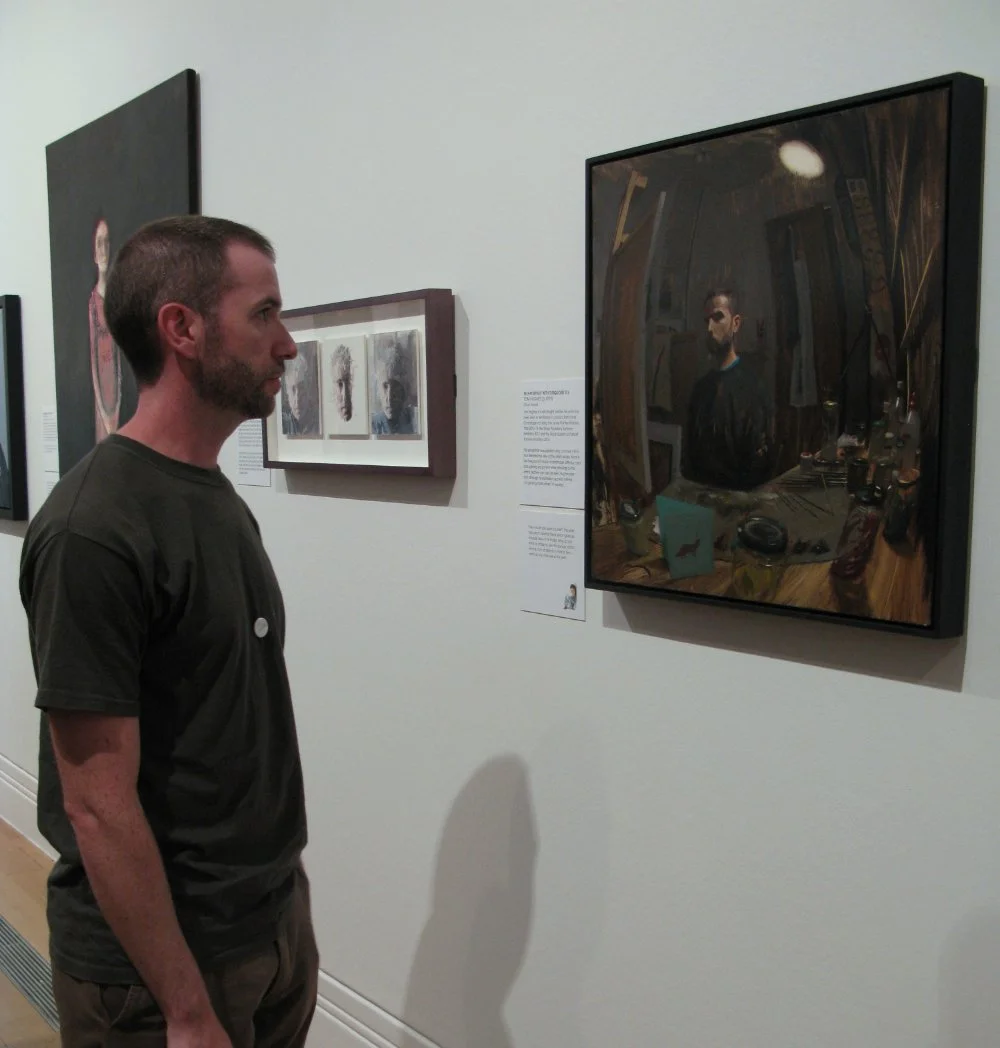 A man with a beard and short hair in a dark t-shirt looks at artwork displayed on a gallery wall, which includes a large, reflective painting of himself and other smaller pieces.
