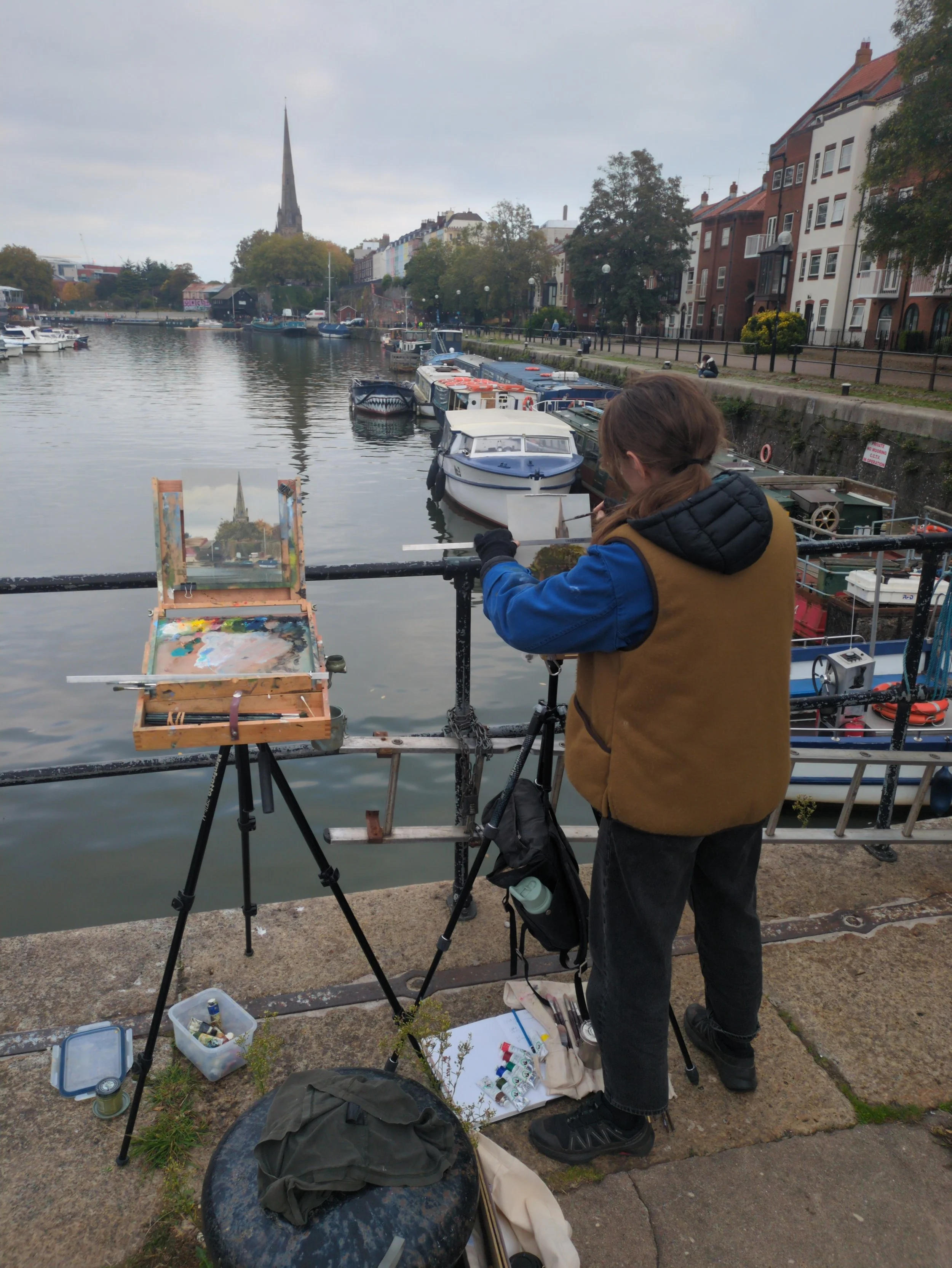 An artist is painting outdoors by a river, with boats docked nearby and a cityscape with a church in the background. They are wearing a brown vest and black gloves, with art supplies around them.