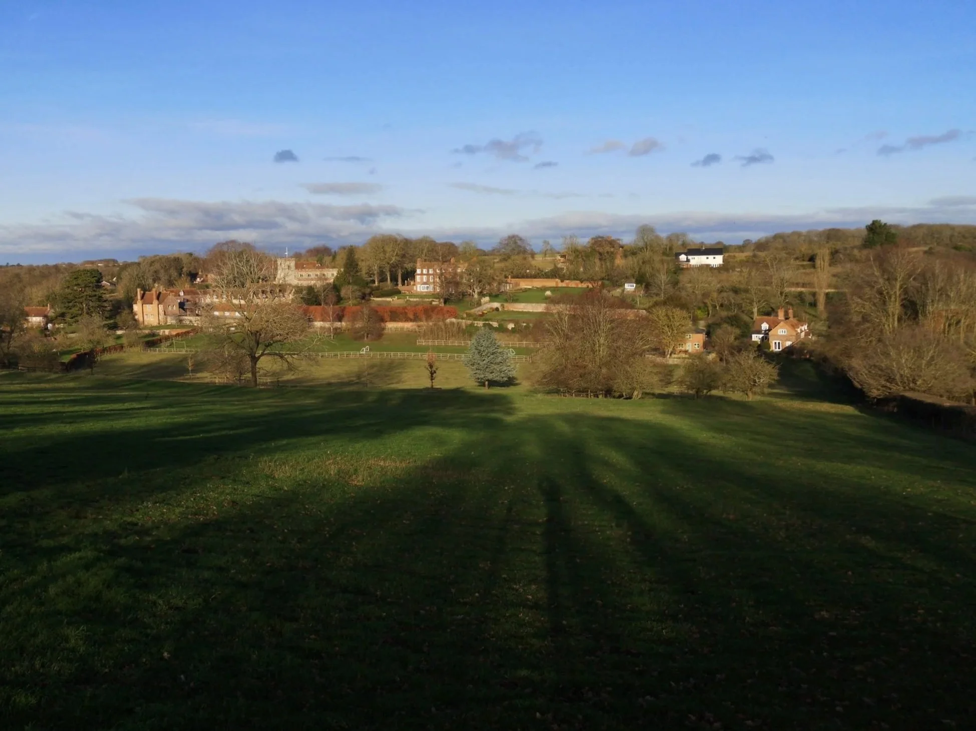 A scenic view of a rural hillside with houses, trees, and green grassy fields under a blue sky with scattered clouds.