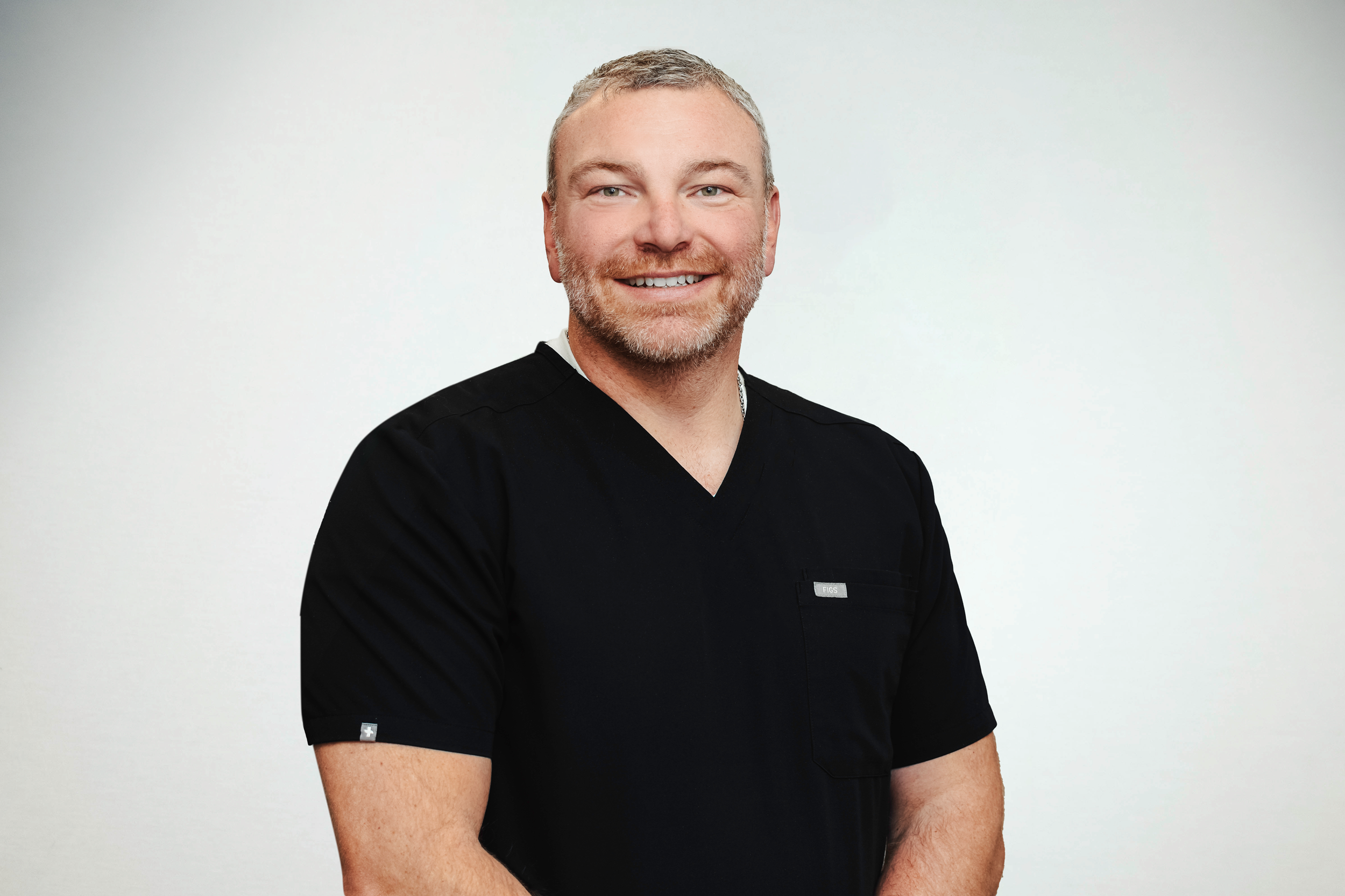 Dr Joshua Vandersteen, wearing black medical scrubs, standing against a plain white background.
