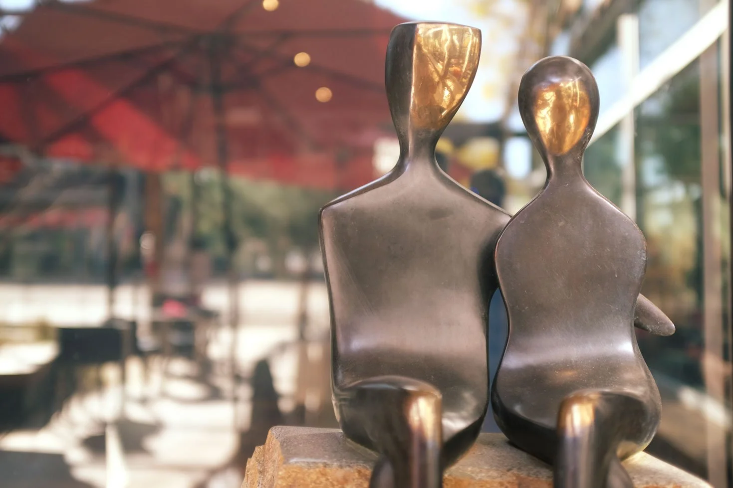 Two abstract metallic sculptures of human figures with simplified features, placed outdoors near a window with reflections of trees and a red umbrella.
