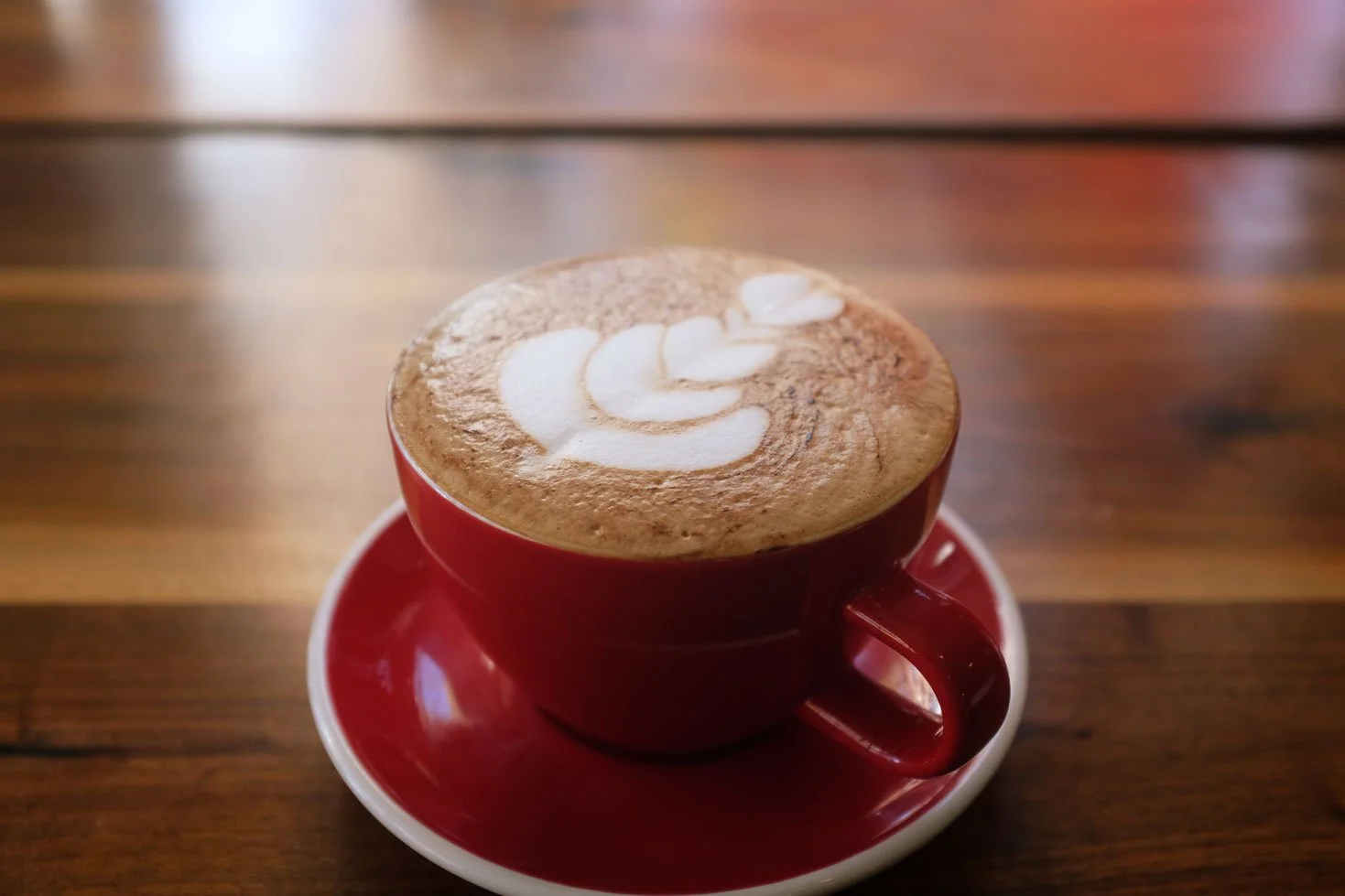 A red coffee cup filled with a cappuccino topped with foam art, placed on a matching red saucer on a wooden table.