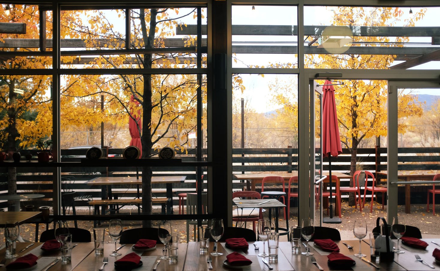 Restaurant interior with tables set with glasses, silverware, and red napkins, looking out through large glass windows to a patio with tables, chairs, red umbrellas, and trees with yellow autumn leaves.