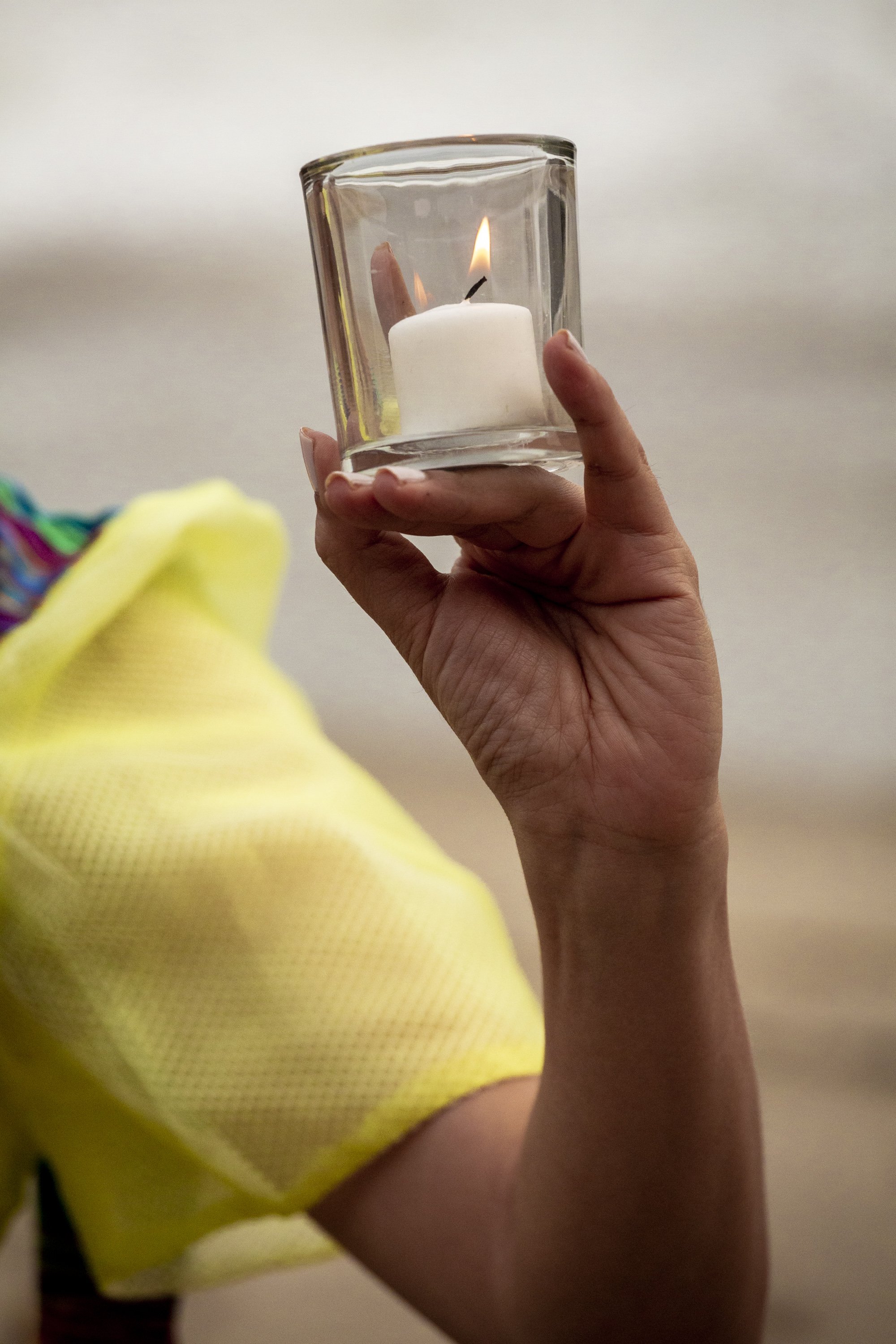 Person holding a clear glass container with a lit white candle inside, the person's hand is visible, and they are wearing a yellow garment.